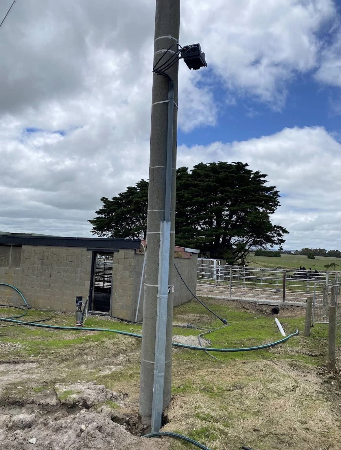 A utility pole with electrical equipment in a rural area with a cloudy sky, a small building, and fenced fields nearby.