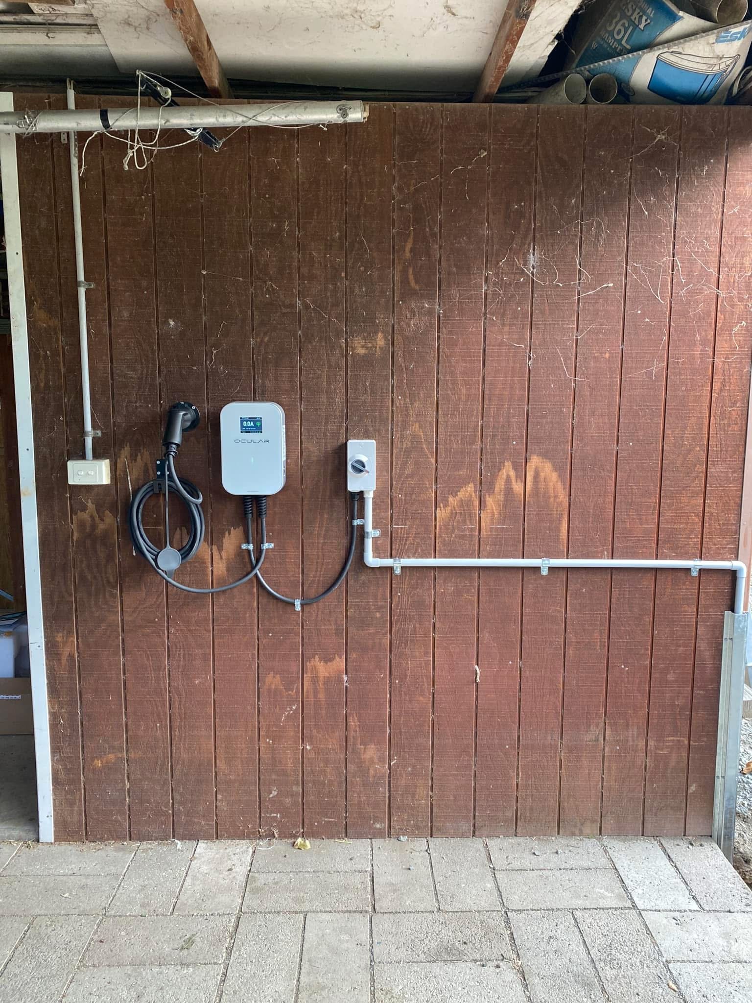 Wooden wall with electrical components and a charging cable attached, along with some pipes and boxes above, and concrete tiles on the ground.
