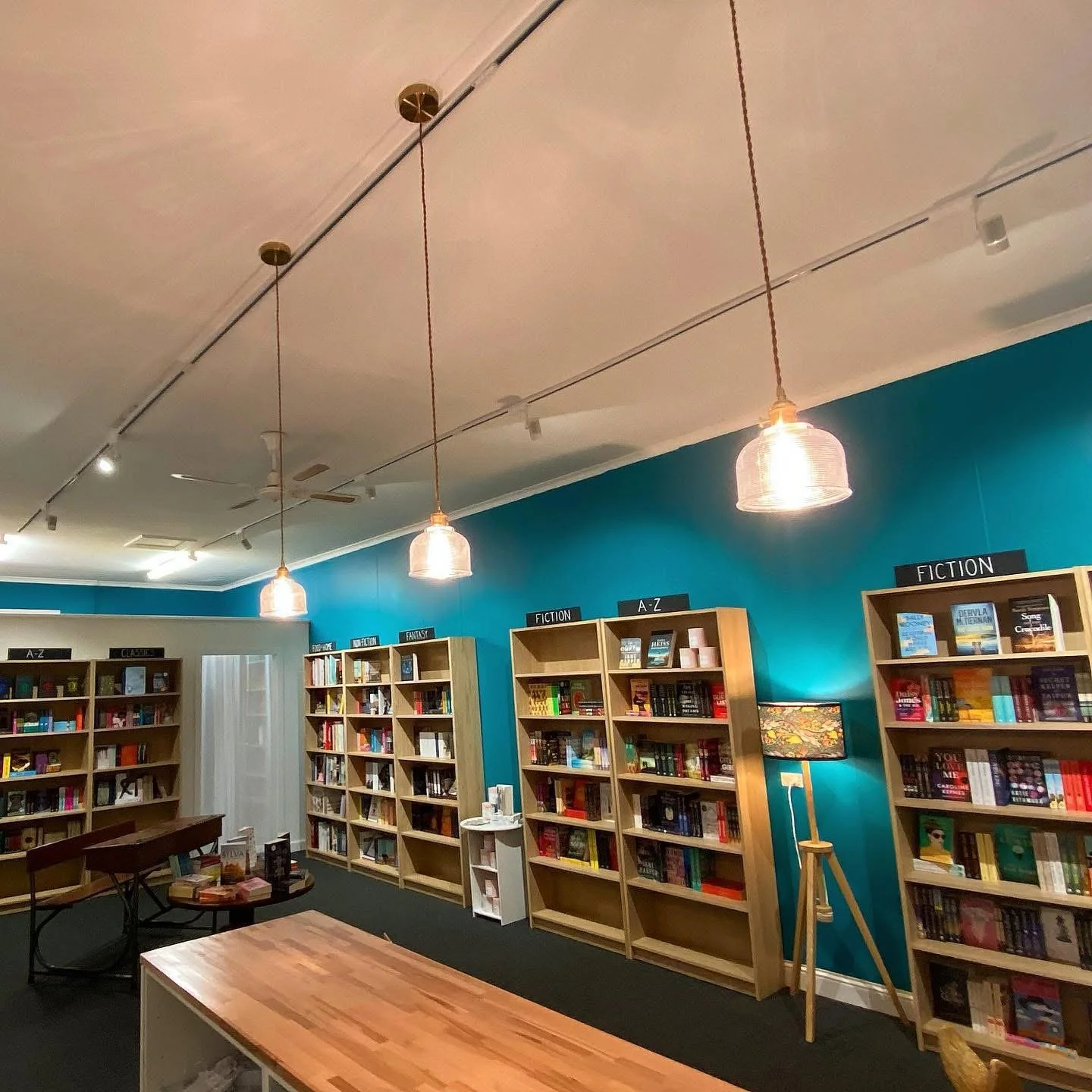 Inside a bookstore with teal walls, wooden bookshelves labeled with genres like Fiction, Fantasy, Non-Fiction, and A-Z, lit by hanging pendant lights and a tall floor lamp, with tables in the foreground.