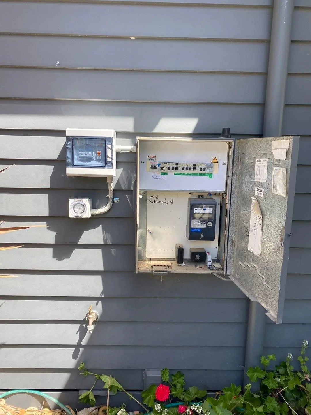 An outdoor electrical meter and breaker panel mounted on a gray house exterior wall with some plants and flowers beneath.