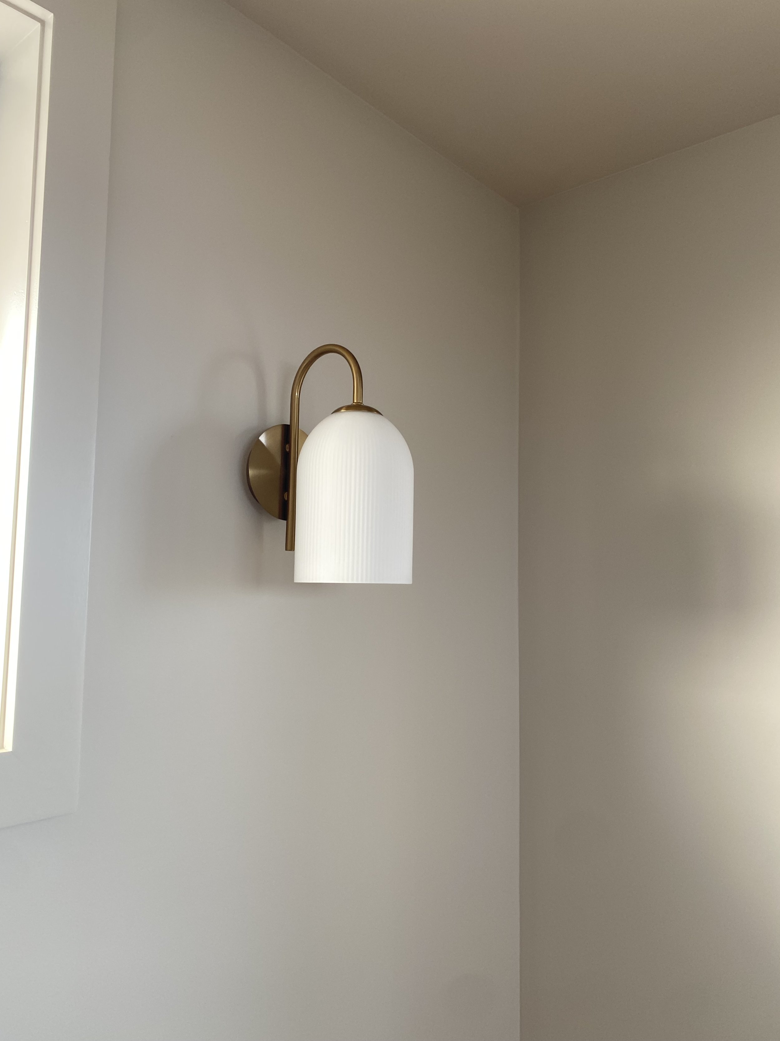 A wall-mounted light fixture with a brass curved arm and a white ribbed glass shade on a plain, light-colored wall.