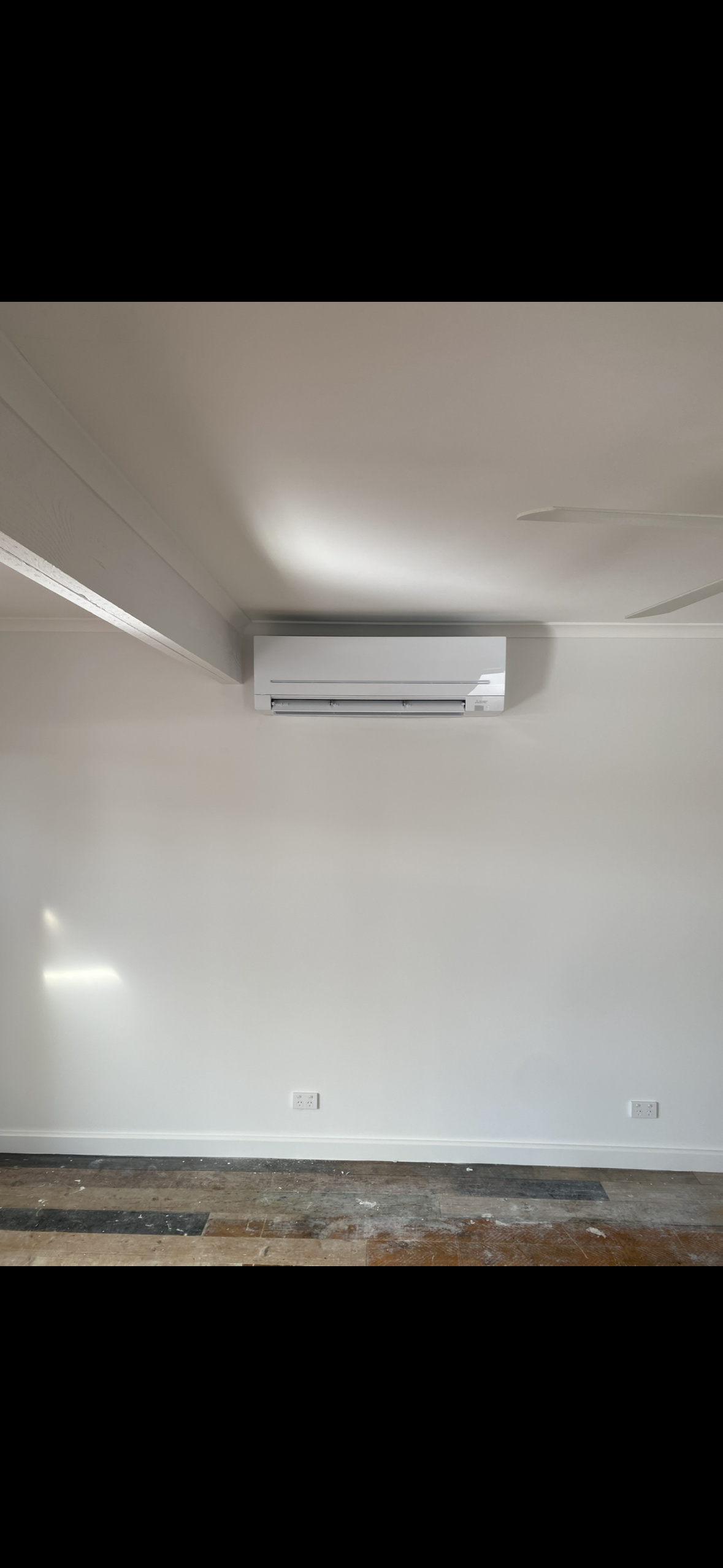 Empty wall with electrical outlets, white air conditioning unit installed high on the wall, and a plain white ceiling. The floor shows some unfinished wood or construction debris.