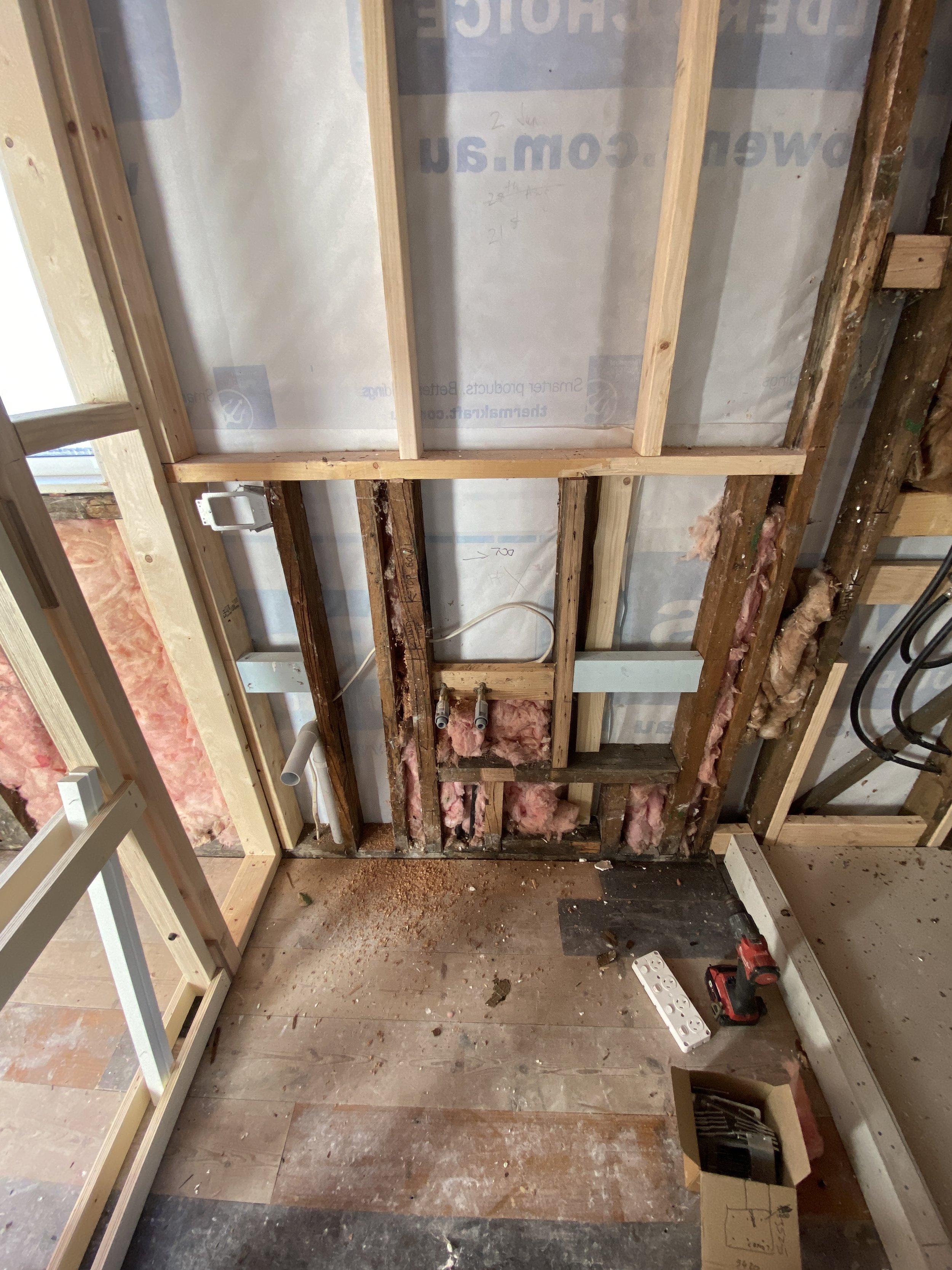Partially removed wall with exposed wooden studs, insulation, electrical wiring, and plumbing, indicating a room renovation or construction process.