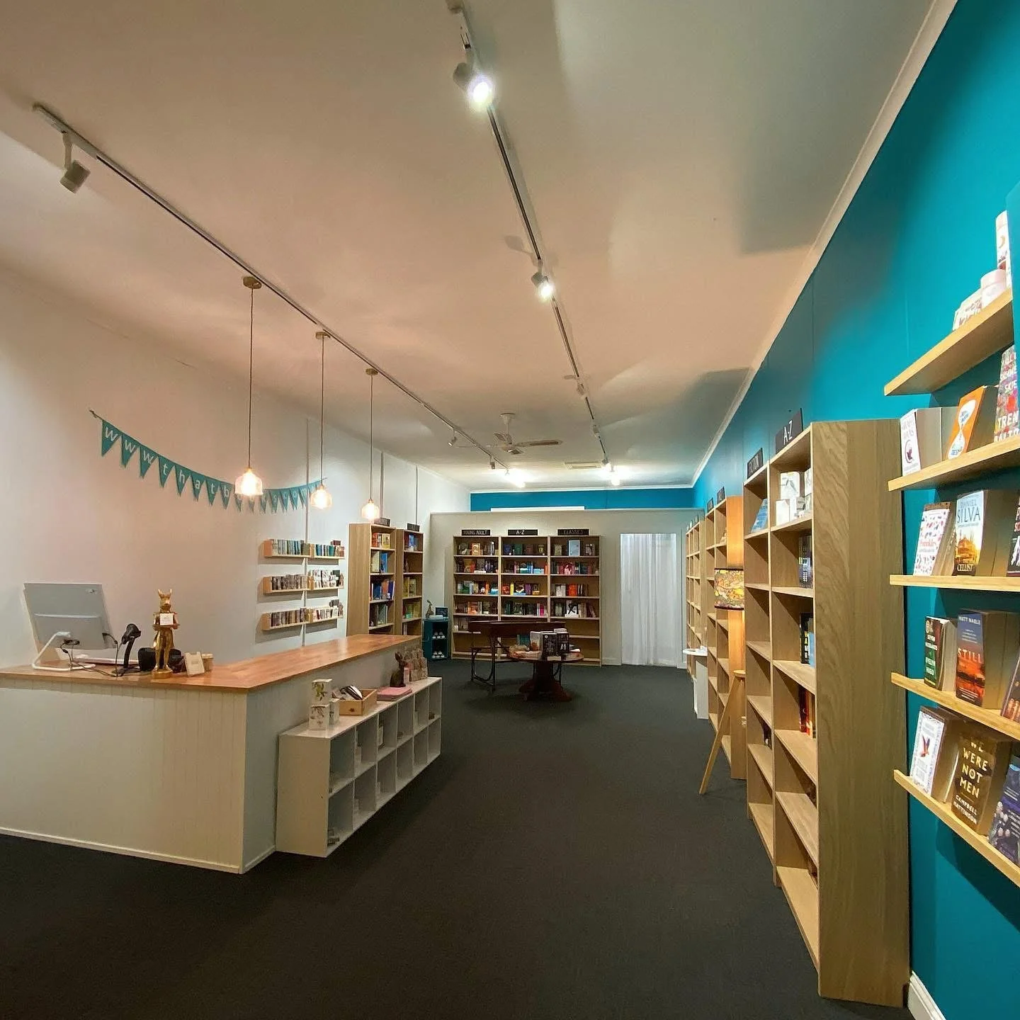 Interior of a bookstore with wooden shelves filled with books, a checkout counter with a computer, decorative items, hanging pendant lights, and a blue accent wall with shelves.