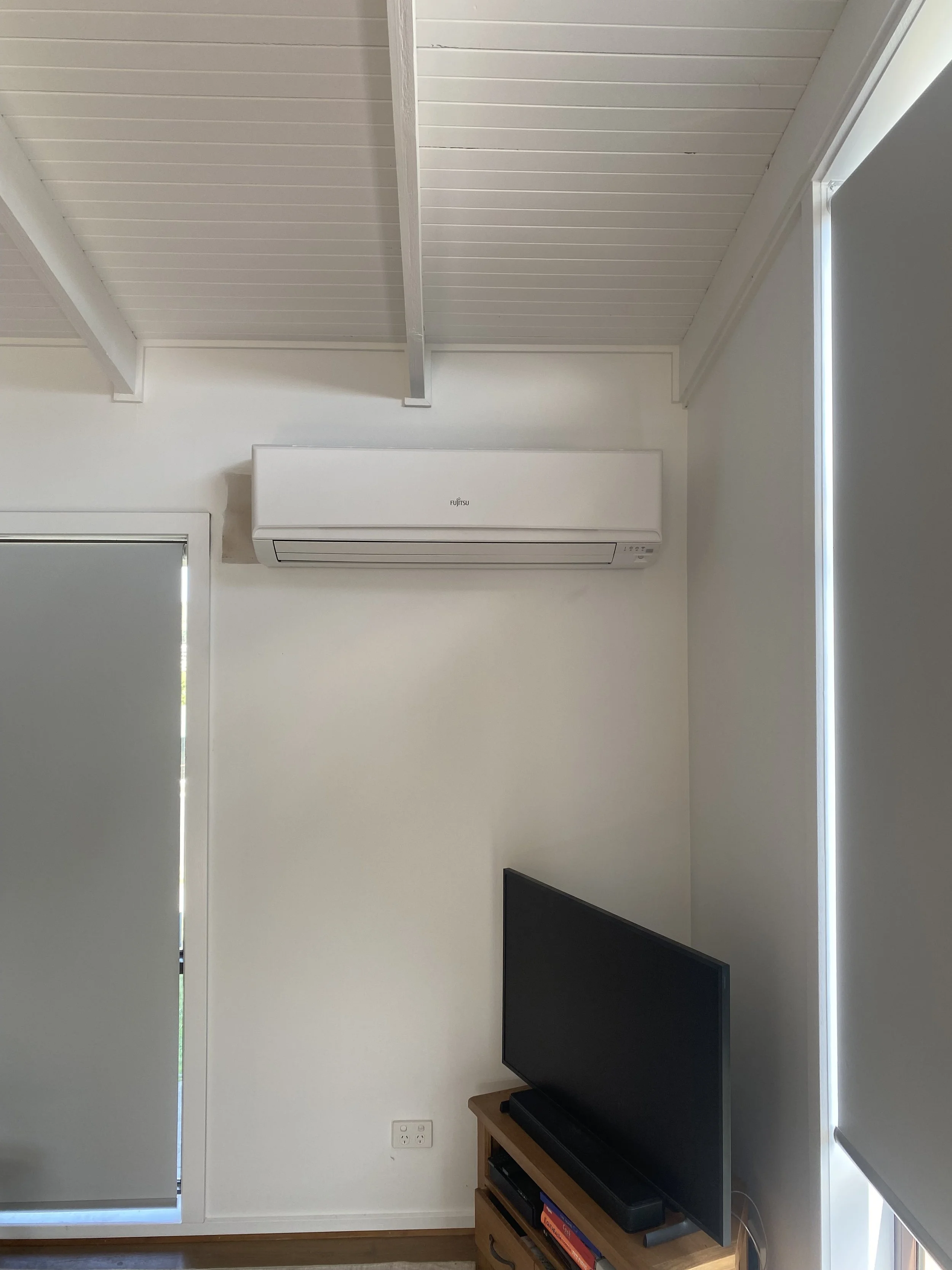 Living room corner with white walls, a heater/air conditioner mounted high on the wall, a window with vertical blinds, and a wooden TV stand with a flat-screen TV and books.