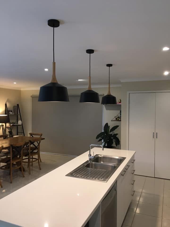 Interior view of a modern kitchen with a white island, stainless steel sink, three black pendant lights, a dining area with wooden chairs, and a beige wall.