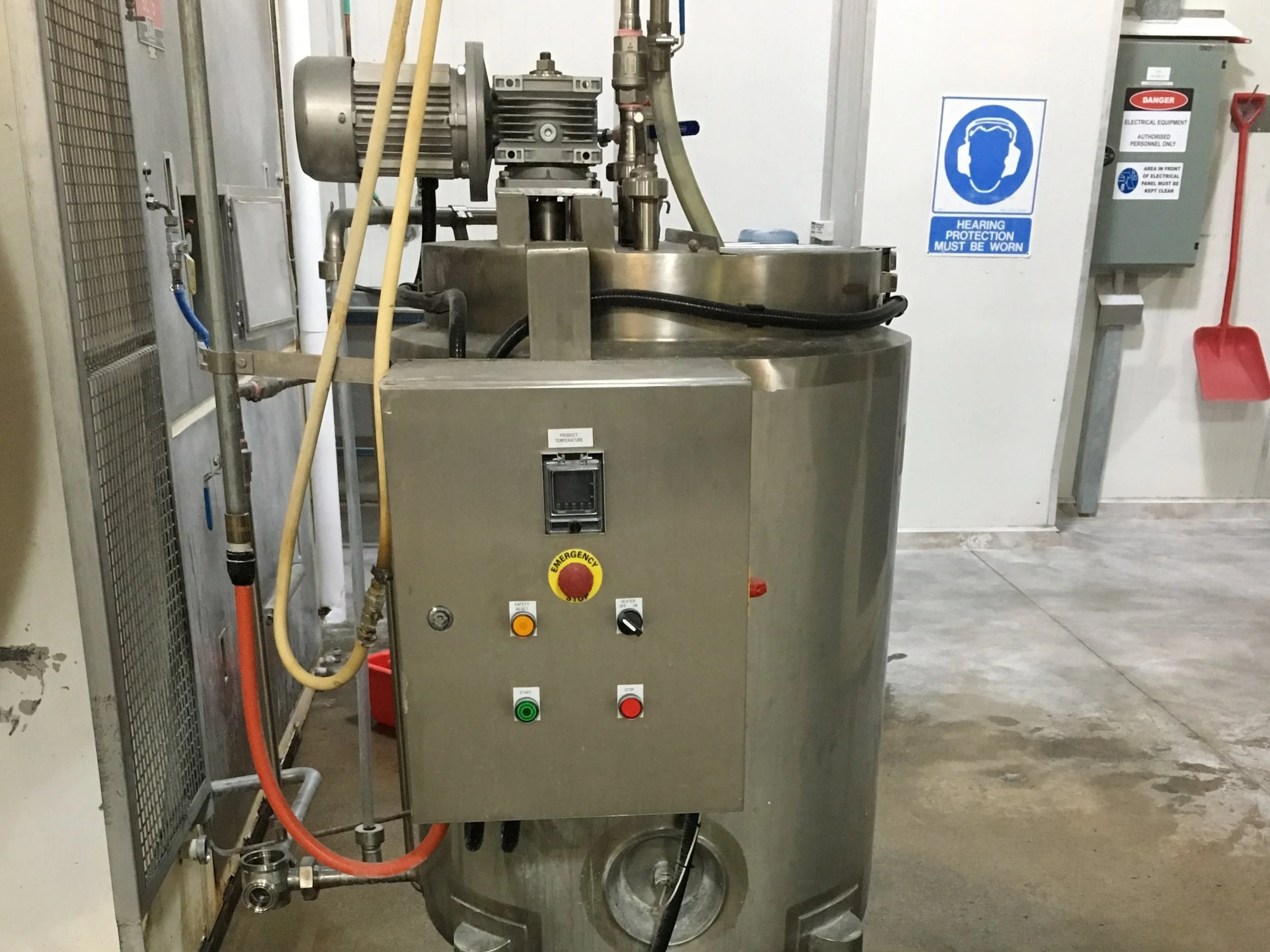 Industrial stainless steel machine with control panel, buttons, and pipes in a mechanical or manufacturing setting. Signs on the wall indicate hearing protection must be worn and electrical safety measures.