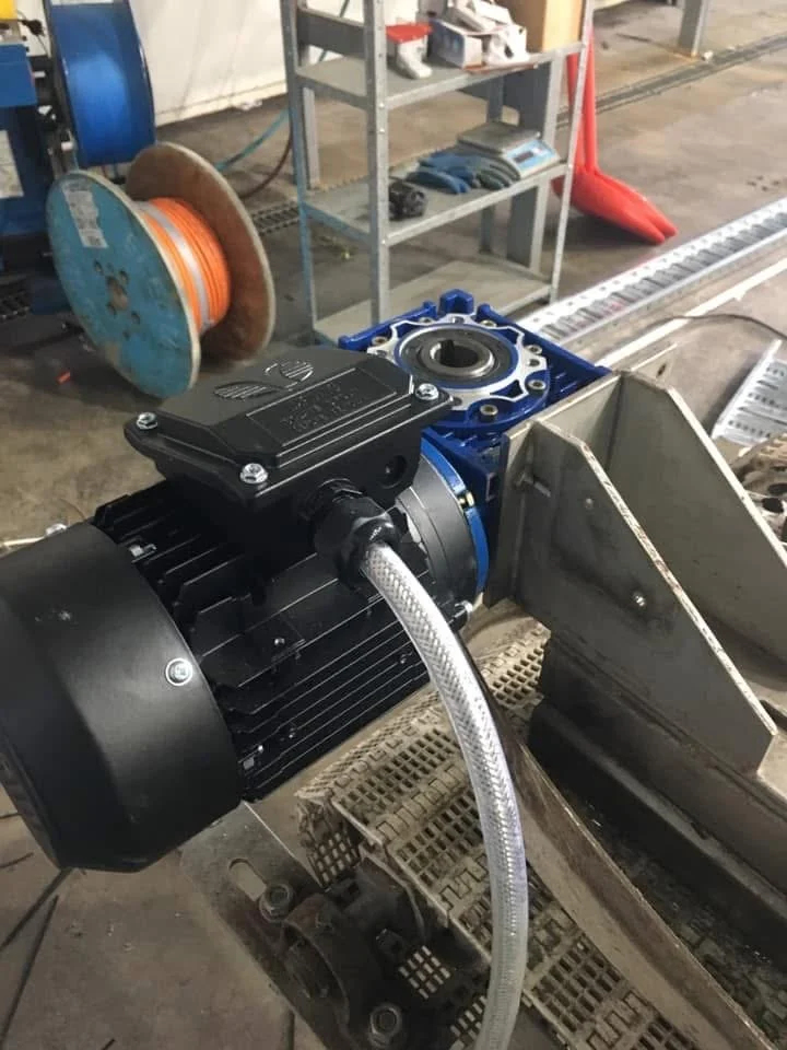 Close-up of an industrial electric motor attached to a blue mechanical component, with a clear braided hose connected to it, in a workshop or manufacturing setting.
