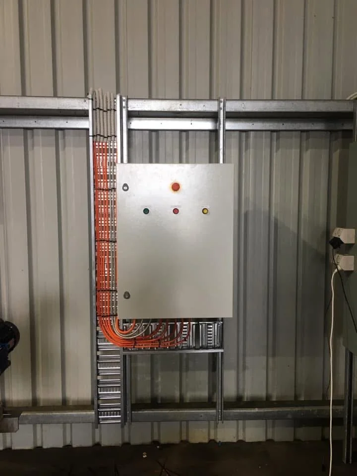 Electrical control panel mounted on a metal frame with wiring and buttons, set against a metal wall in an industrial setting.