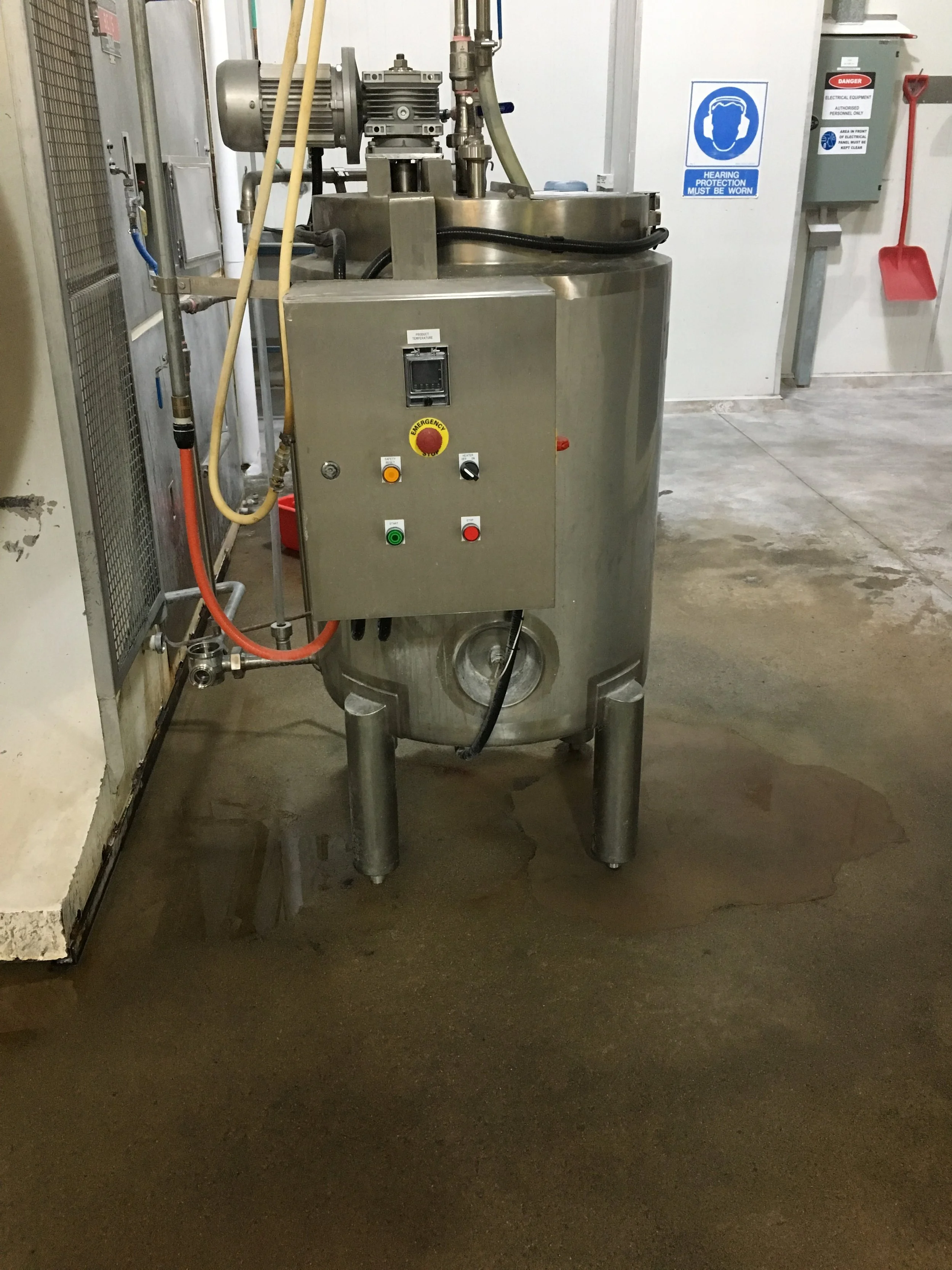 Stainless steel industrial machine in a room with safety signs on the wall, including hearing protection and electrical equipment warnings, and a red snow shovel hanging on the wall.