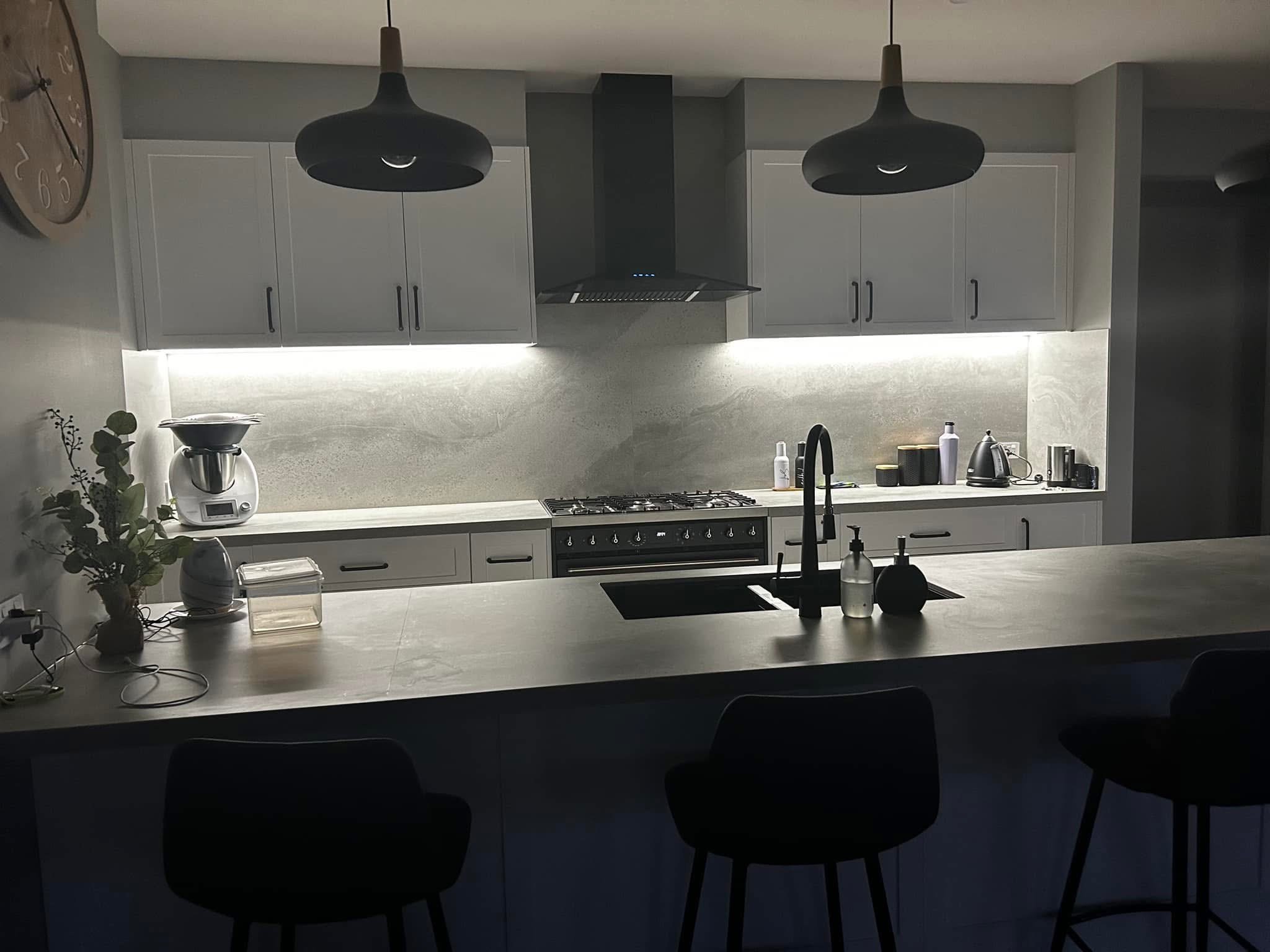 Modern kitchen with white cabinetry, a black range hood, and black pendant lights above the island. The countertop has a plant, a mixer, and kitchen appliances. The backsplash and walls are in light neutral tones, with under-cabinet lighting illumina