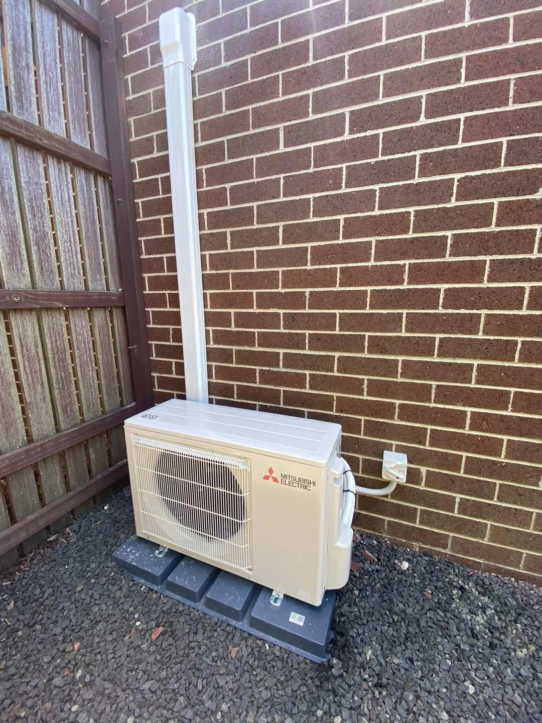 An outdoor air conditioning unit from Mitsubishi Electric installed against a brick wall, with a vertical white PVC pipe nearby and a small wooden fence on the side.