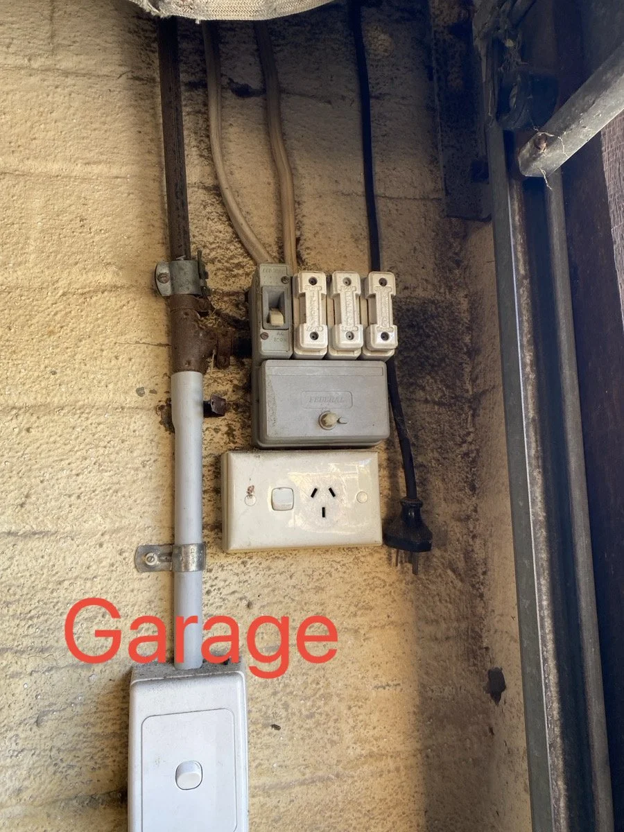 Electrical meter and switches on a beige stucco wall in a garage, with electrical cords and conduit visible.