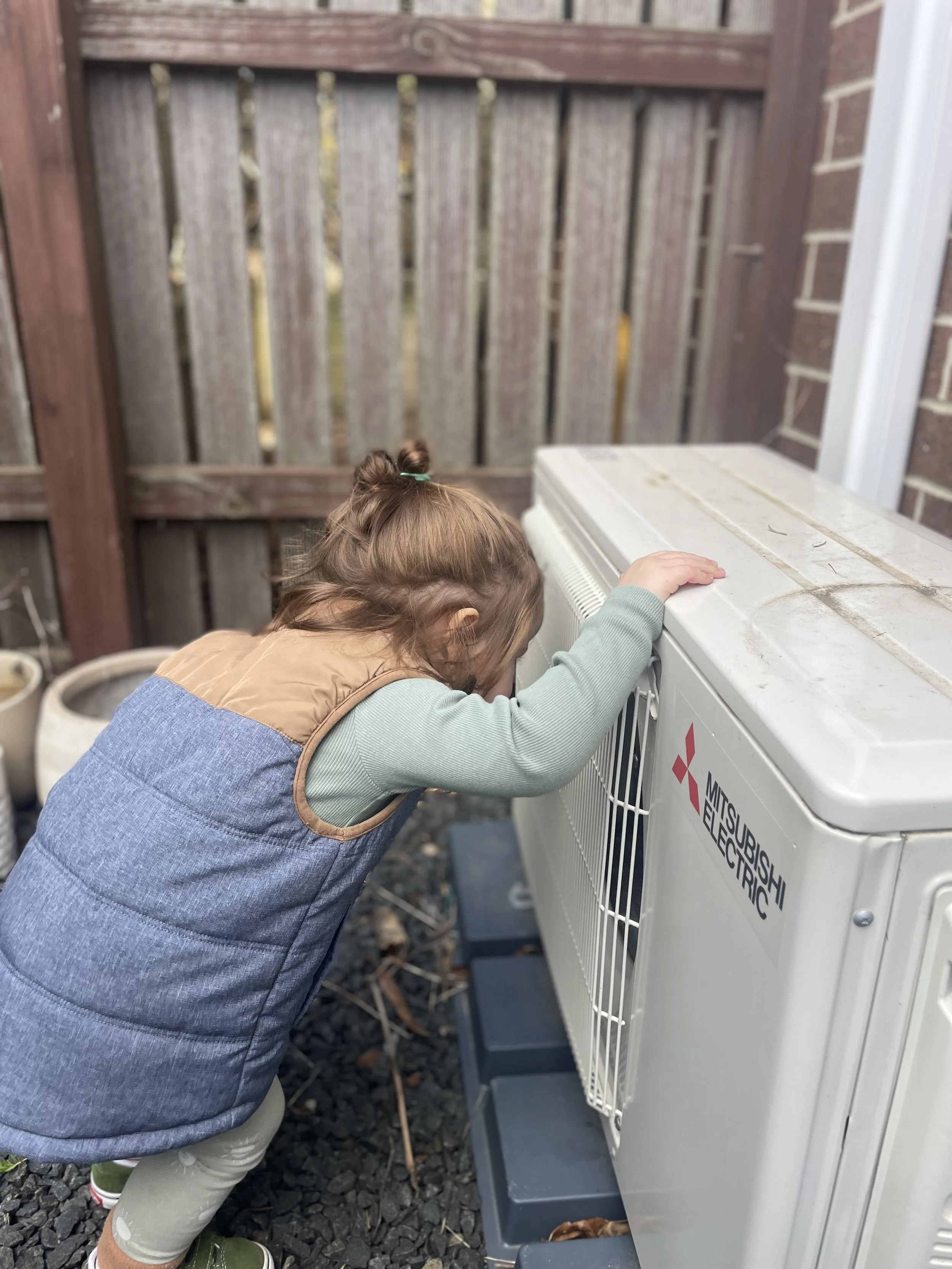 A young girl with a topknot hairstyle is leaning over to look inside an electrical box or cage mounted outside on a wall, with a wooden fence in the background.