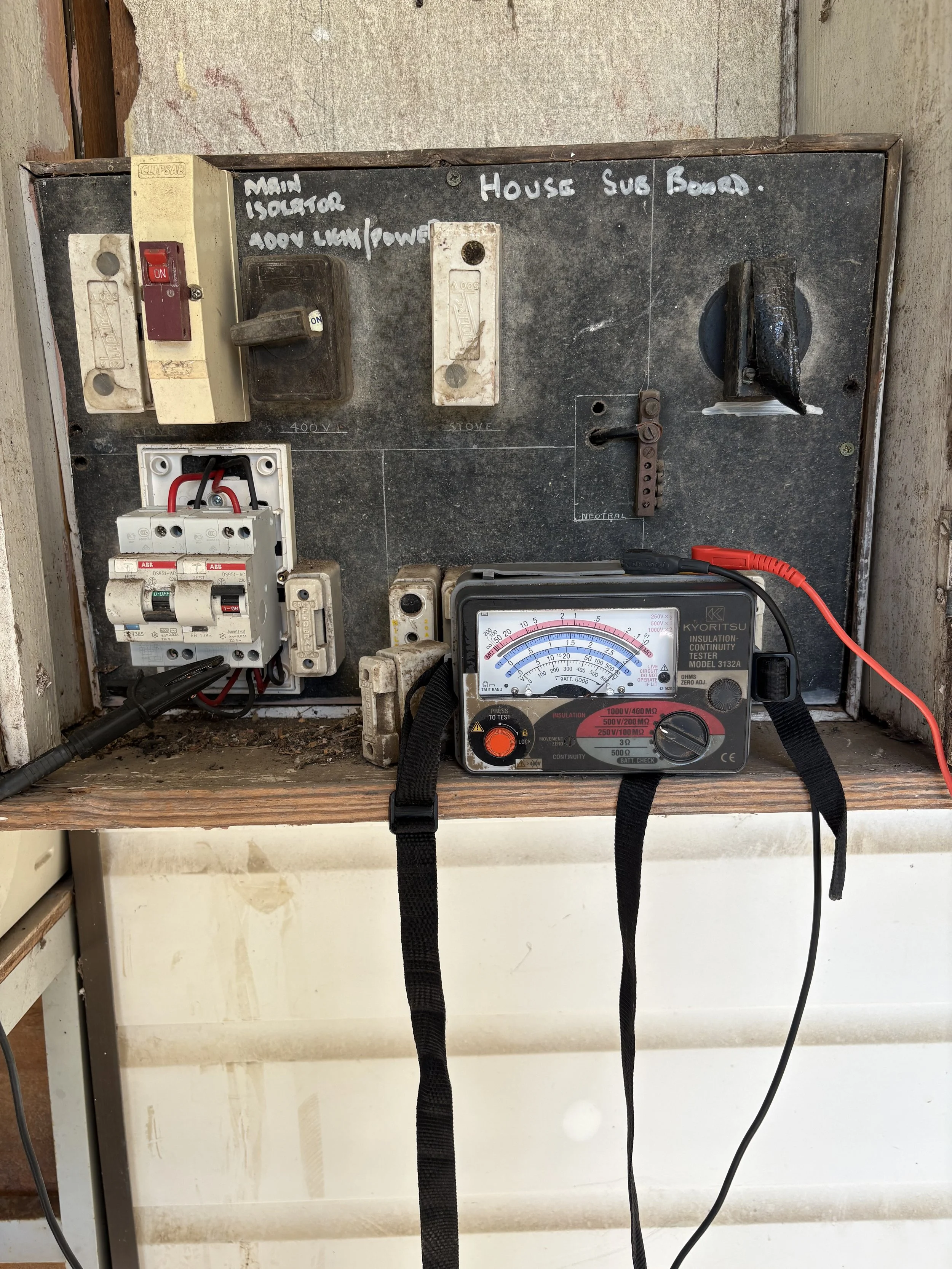 Electrical panel box with circuit breakers, a multimeter, and handwritten labels. The handwritten notes include 'Main Isolator 100A Main Power' and 'House Sub Board'. The multimeter is showing electrical readings and is connected to the panel via bla