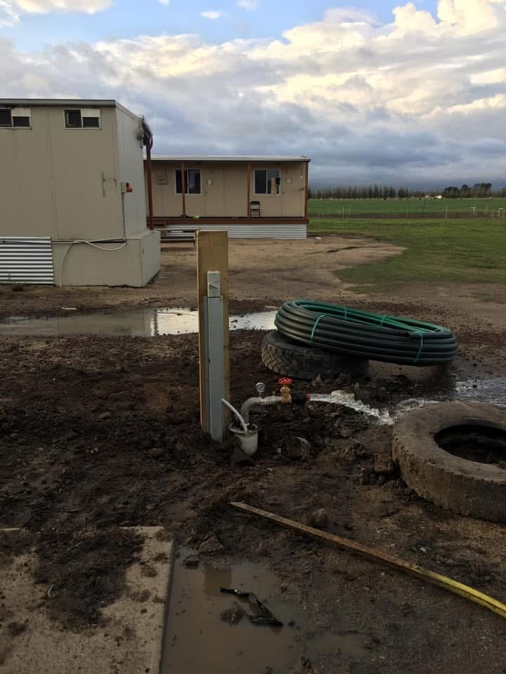Construction site with utility pipes and a frame for an outdoor faucet or water connection, a large tire, concrete drain, and a mobile home in the background under a cloudy sky.