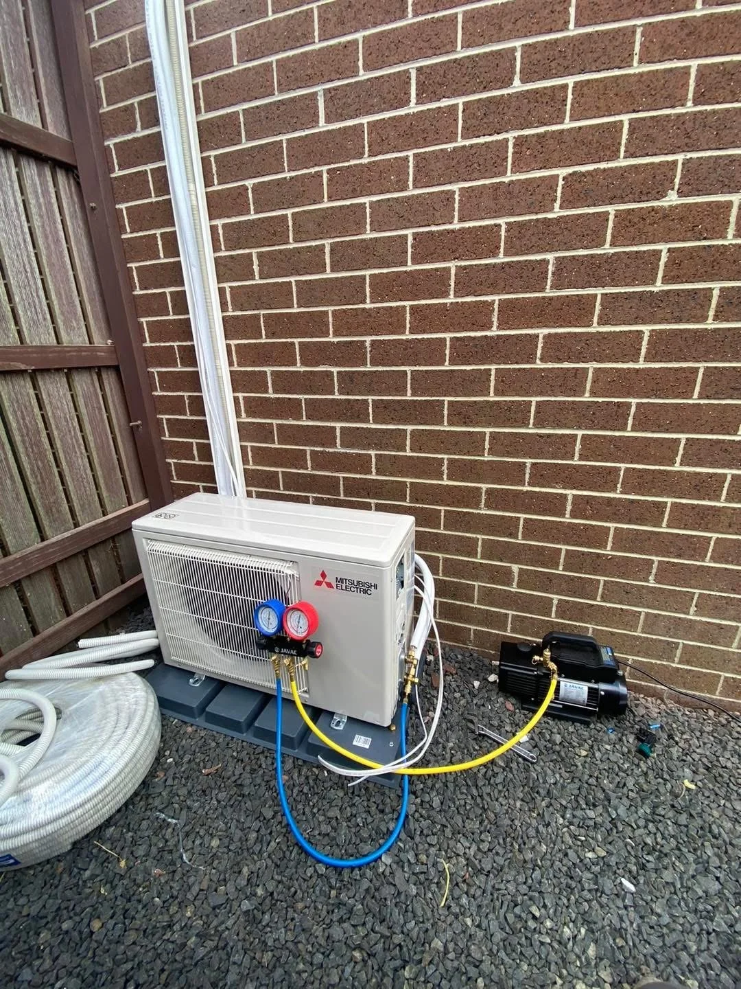 Outdoor air conditioning or refrigeration unit with gauges and hoses connected, situated on a gravel surface against a brick wall, with some piping and tools nearby.