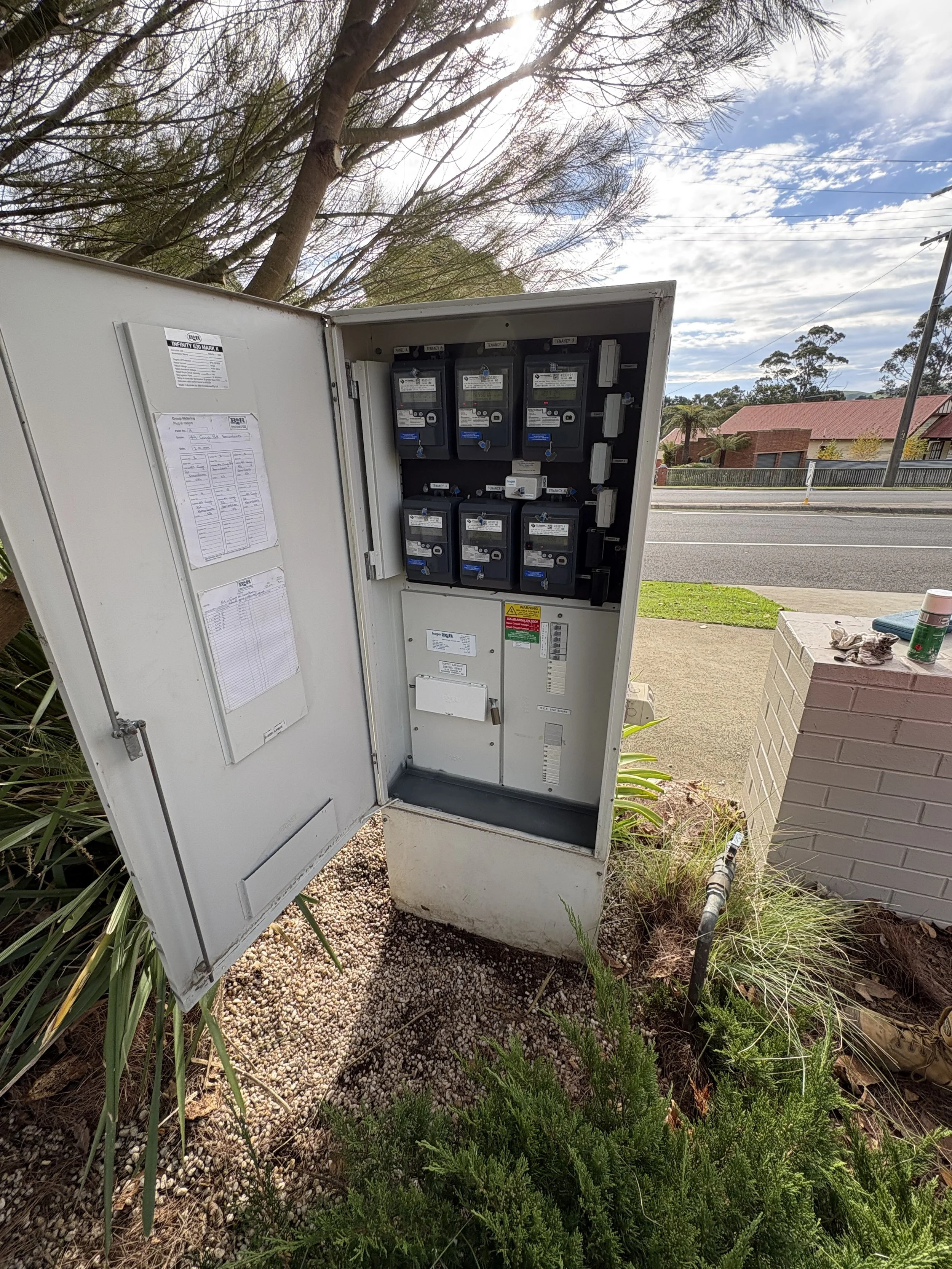 Open electrical meter box on a grassy area with trees and houses in the background, under partly cloudy sky.