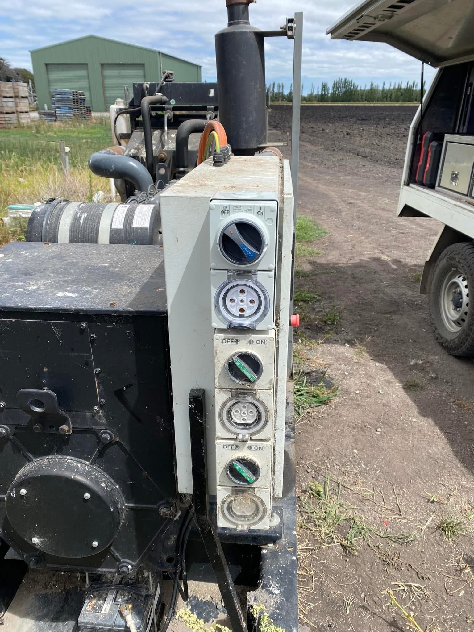 Close-up view of a portable generator with multiple outlets and switches, situated outdoors on a dirt surface with a field and a green building in the background.