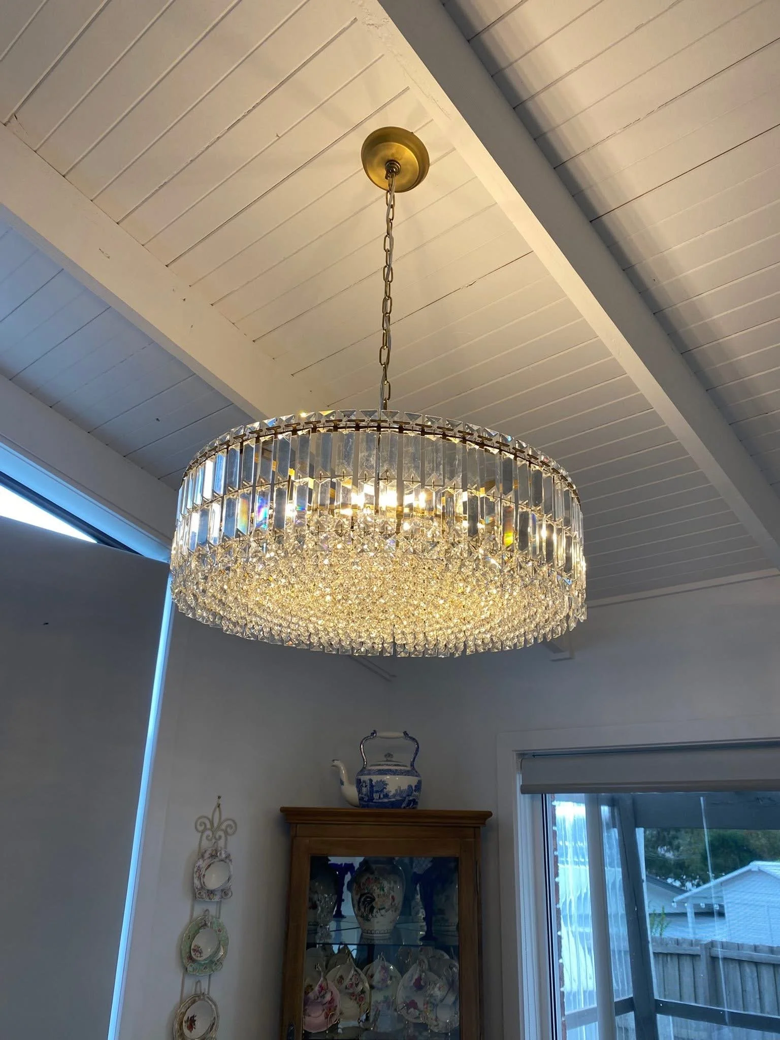 A chandelier with crystal accents hanging from a chain attached to a ceiling with white wooden panels, in a room with a china cabinet displaying decorative ceramics, and a window showing a backyard with a wooden fence and a deck.