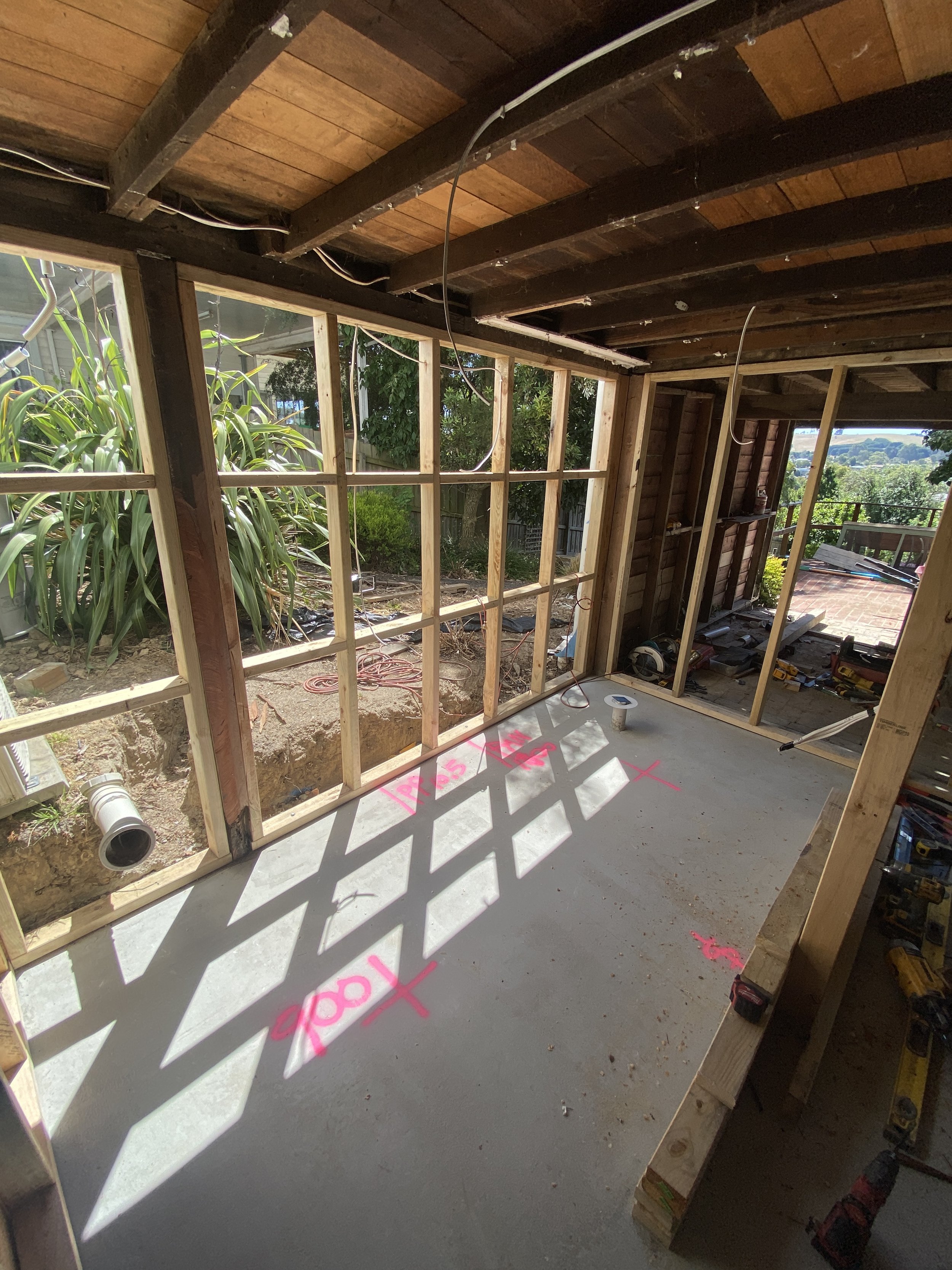Interior view of a house under construction with exposed wooden framing, wiring, and tools on the concrete floor. Outside, there are trees and a brick patio.
