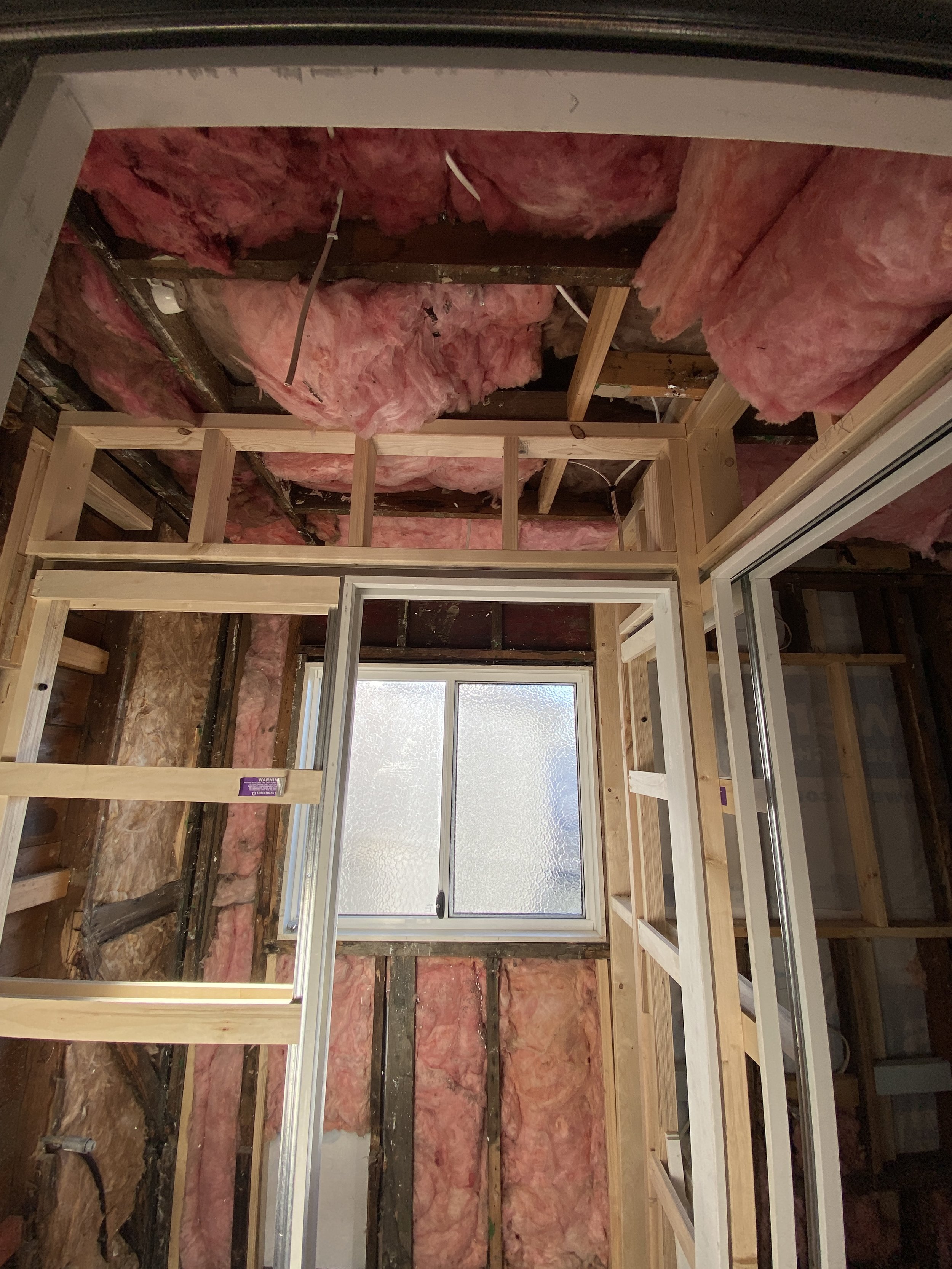 Construction framing in progress with wooden studs, pink insulation, electrical wires, and a window in the background.