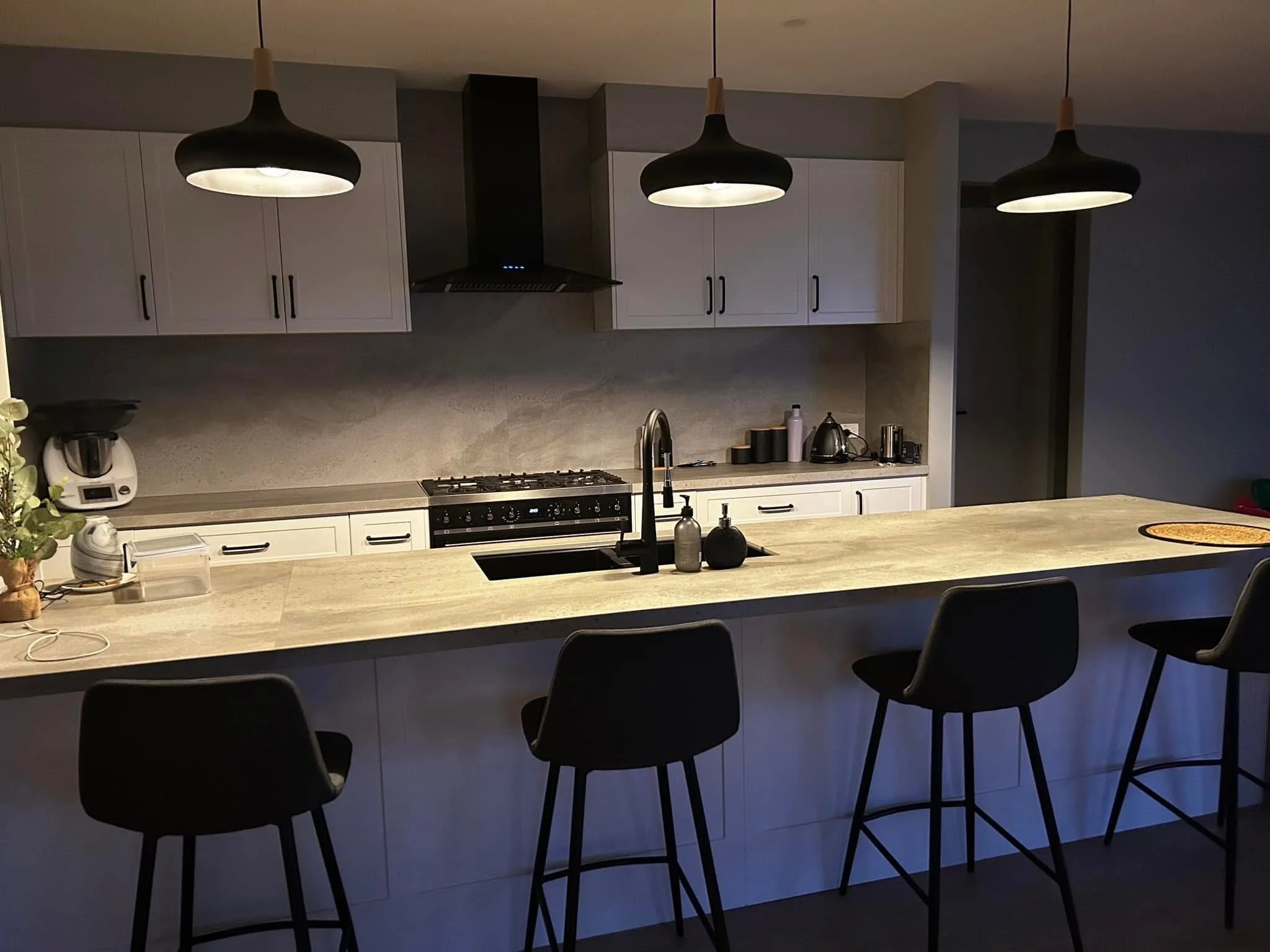 Modern kitchen with four black pendant lights hanging above a light-colored island counter. White cabinets with black handles, a stove with an exhaust vent, a kettle, and various small appliances are visible on the counter. There are four black barst