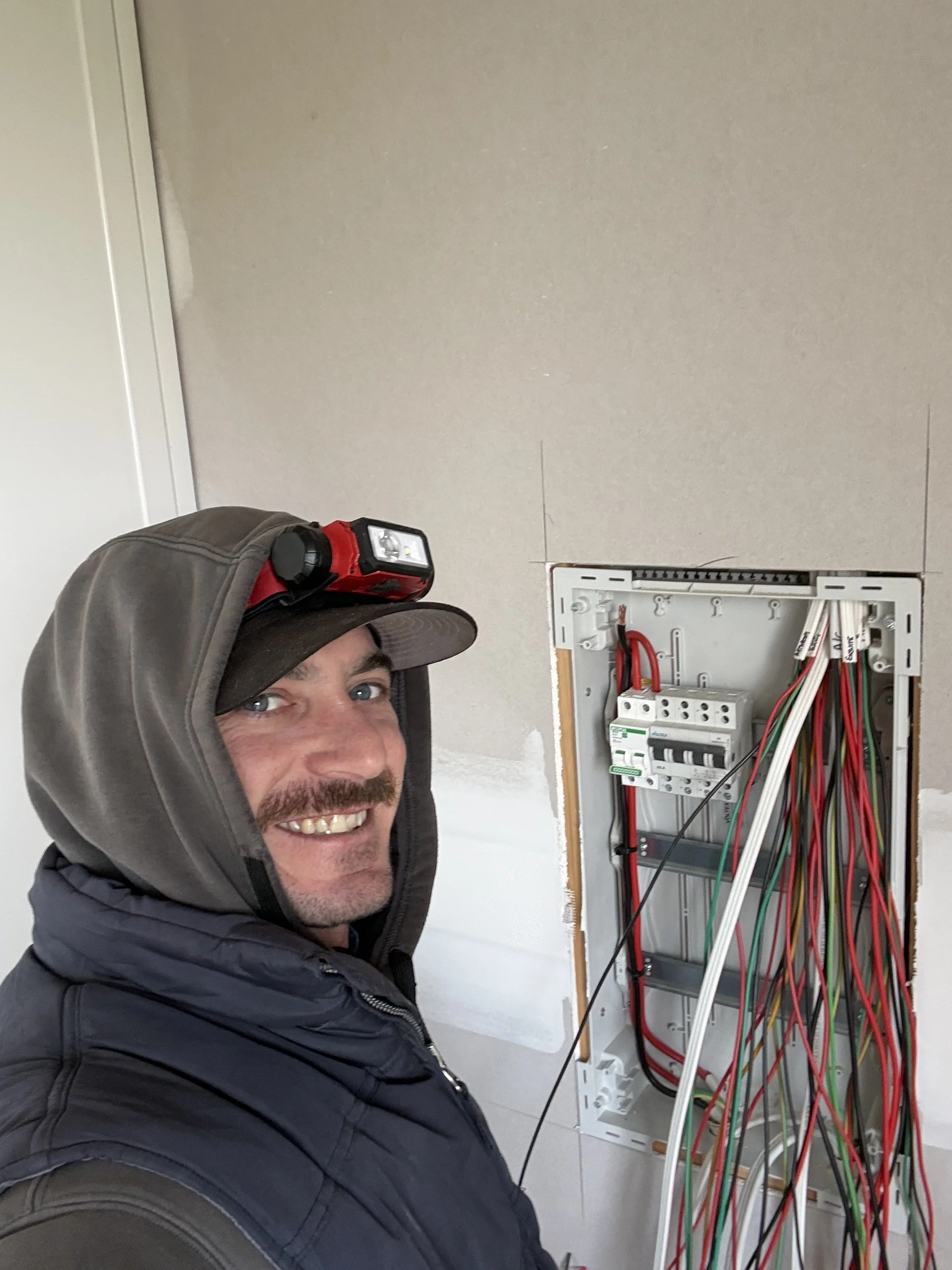 Man in a hoodie and cap with a headlamp smiling at the camera while working on an electrical panel with wires.