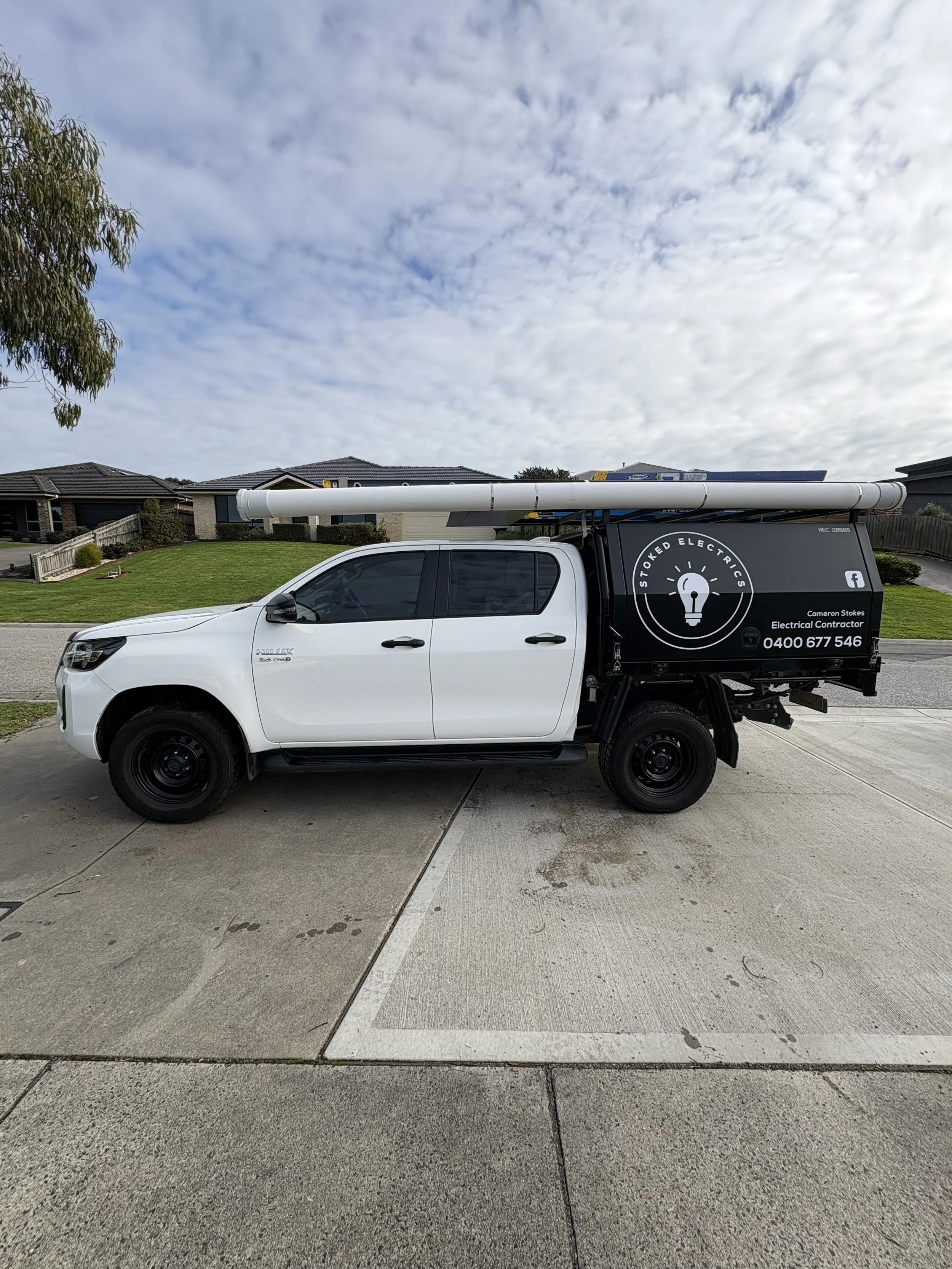 White utility truck with black cargo area, parked on driveway in front of suburban houses, with equipment on roof and signage for an electrical contractor.