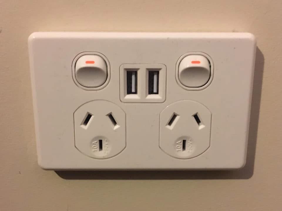 A white electrical outlet with two sockets, two switches, and two USB ports on a beige wall.
