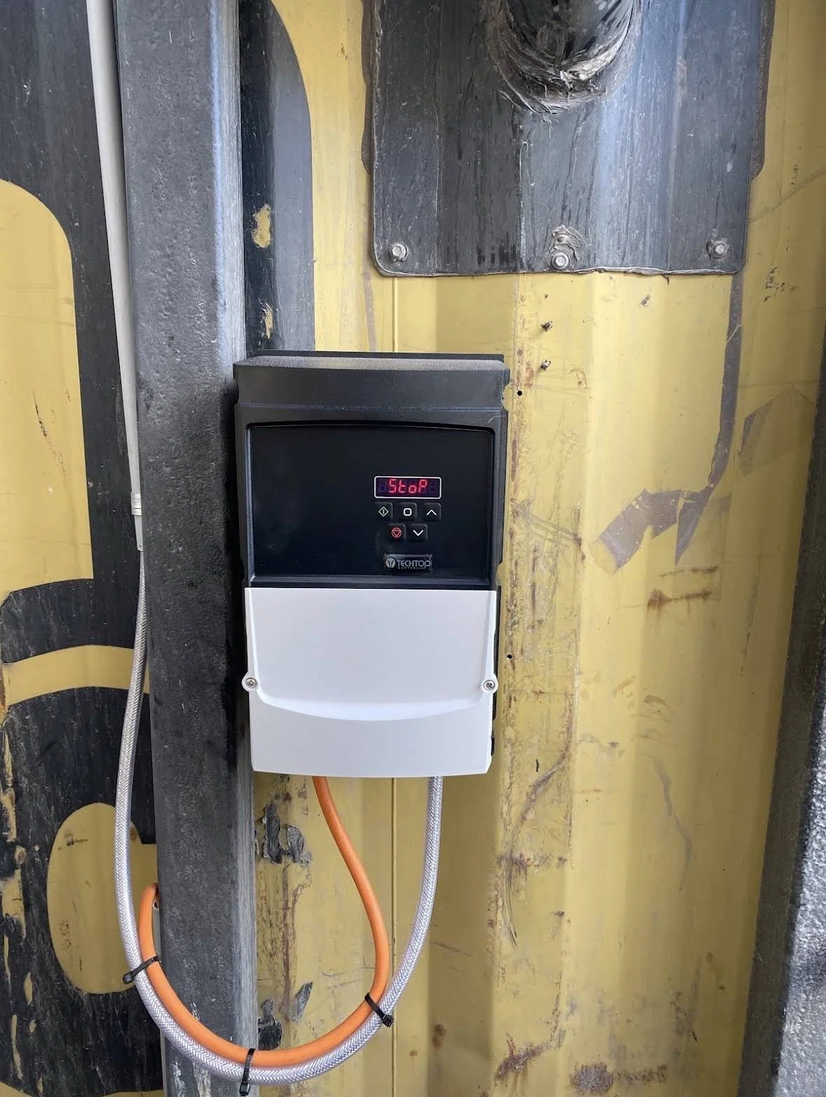 Electric vehicle charging station with a digital display showing 'Stop', connected to cables and mounted on a wall with yellow paint and exposed steel framing.