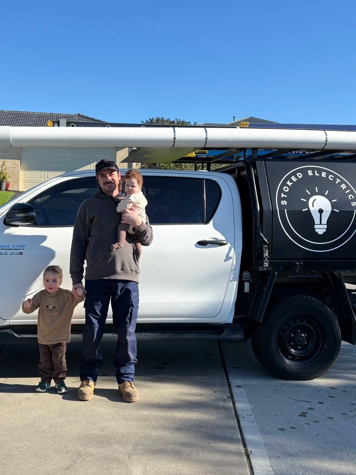 Family standing in front of a white pickup truck with a logo for stoked electric on the side. The father is holding a baby, and a young boy is holding his hand. The background shows a suburban street and houses.