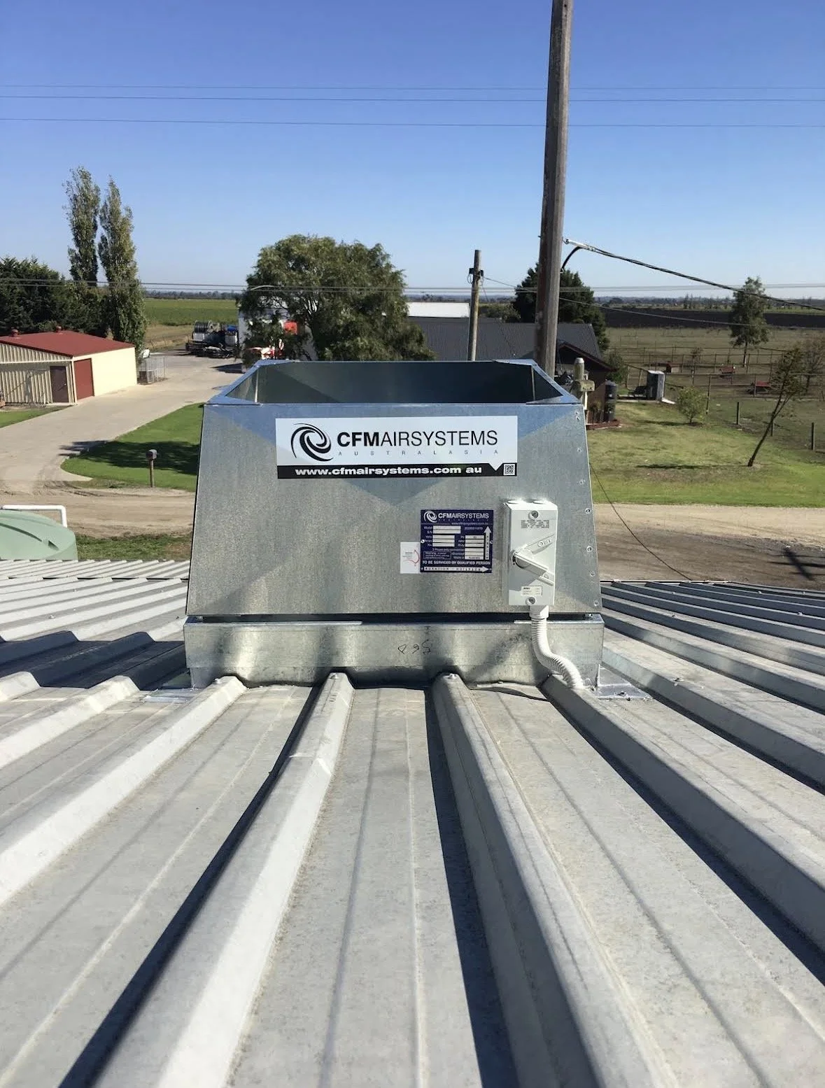 Metal roof with a ventilation fan unit labeled 'CFM AIR SYSTEMS Australia' and a website link, set against a rural landscape with trees, a dirt road, and utility poles under a clear blue sky.
