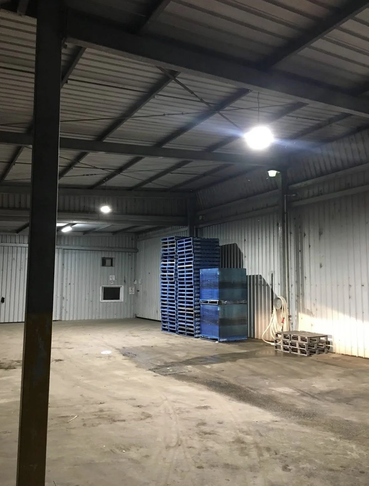 Interior of an industrial warehouse with metal walls and ceiling, illuminated by bright overhead lights, with stacks of blue pallets and a hose reels on the wall.