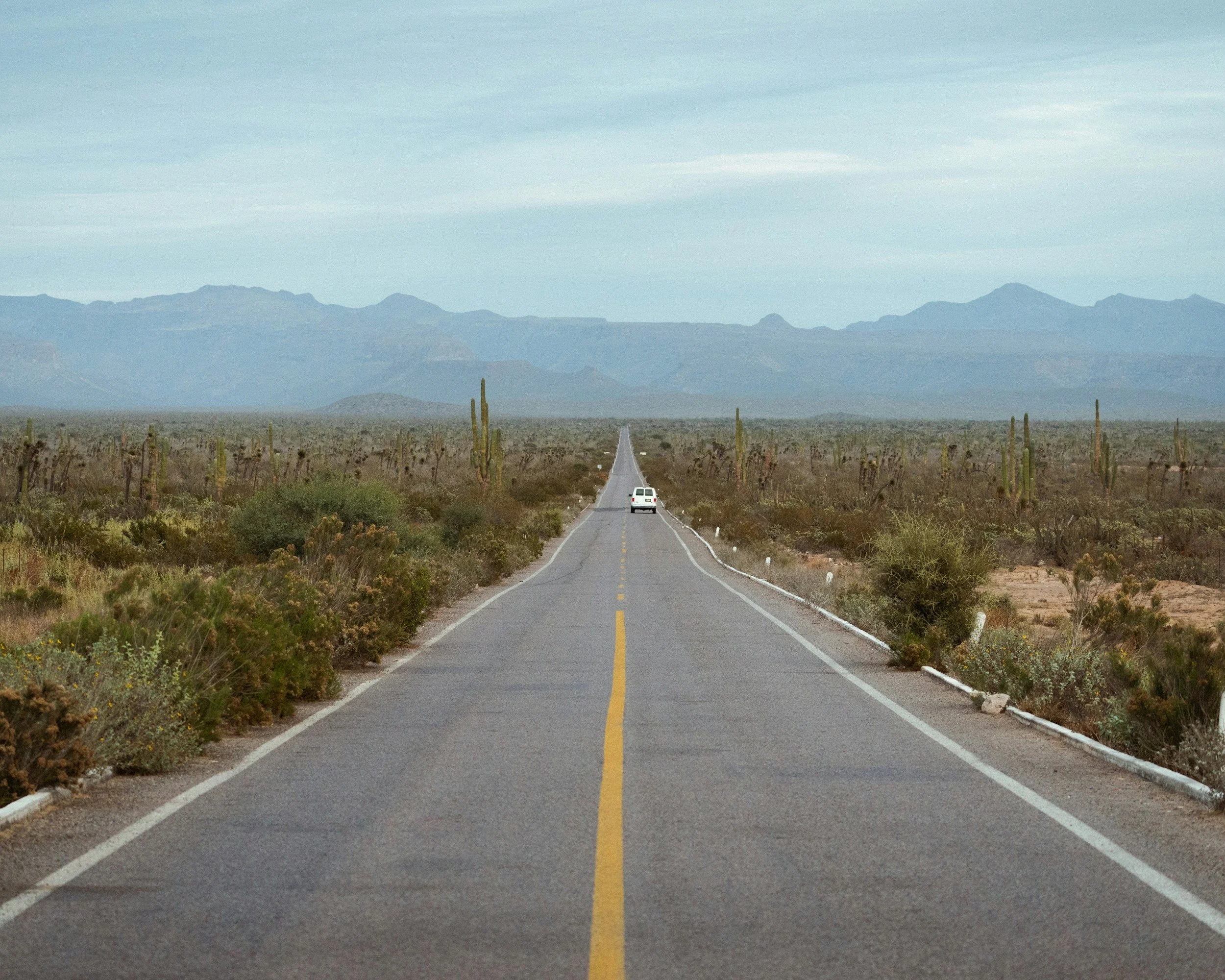 Long straight desert road with a single white car driving away towards mountains in the distance under a cloudy sky.