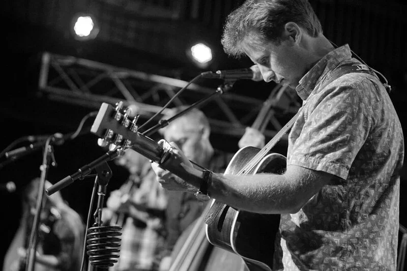 A man playing acoustic guitar on stage during a live performance, with microphones and stage lighting visible.