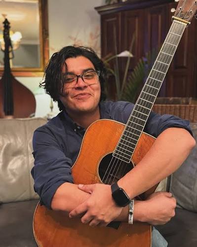A man with curly black hair and glasses sitting on a couch, holding an acoustic guitar, smiling in a living room with wooden furniture and a mirror on the wall.