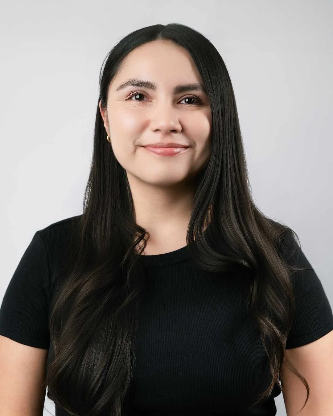Portrait of a woman with long dark hair, wearing a black shirt, smiling, against a plain light gray background.