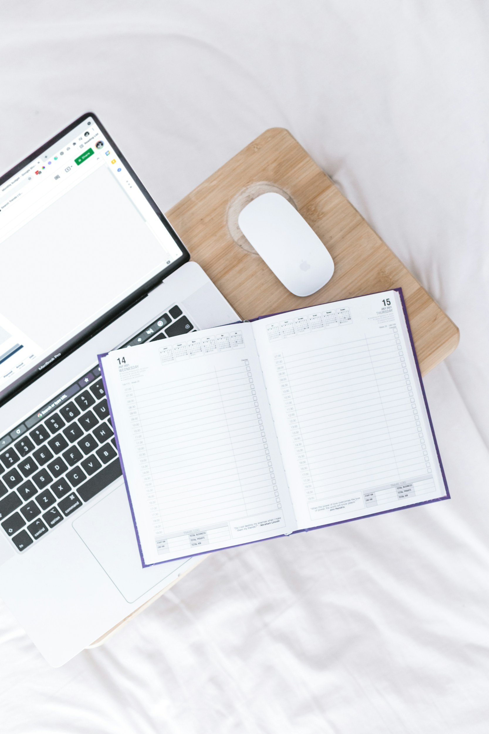 A laptop, a white computer mouse, an open planner, and a wooden desk are arranged on a white surface.