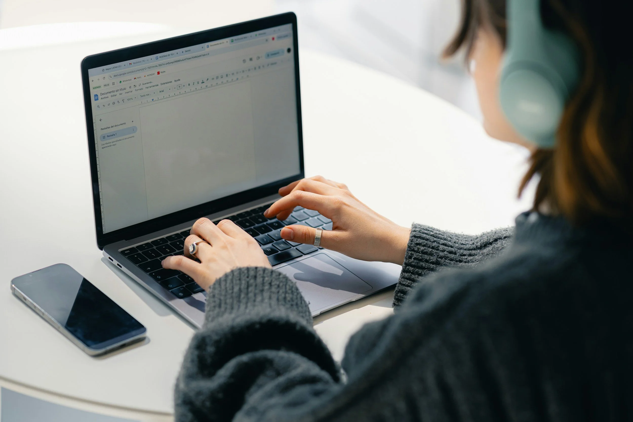 Virtual assistant working on a laptop with a smartphone and tablet on a white table, wearing a face mask.