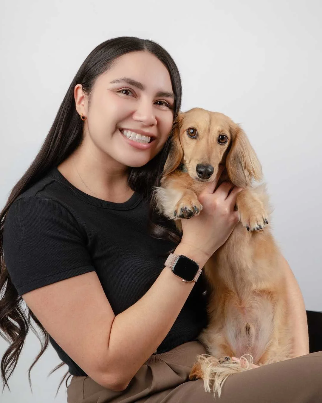 Young woman with long dark hair smiling and holding a light brown long-haired Dachshund puppy against a plain white background.