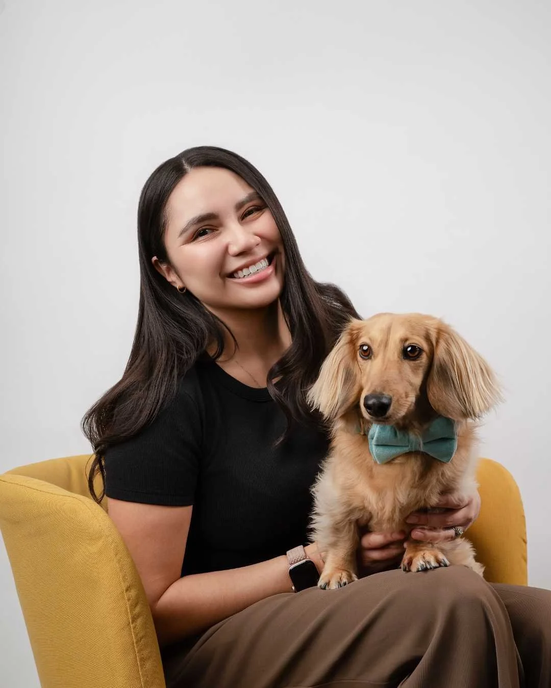 A woman with long dark hair and a black shirt sitting on a yellow chair, smiling and holding a long-haired dachshund dog wearing a green bow tie.