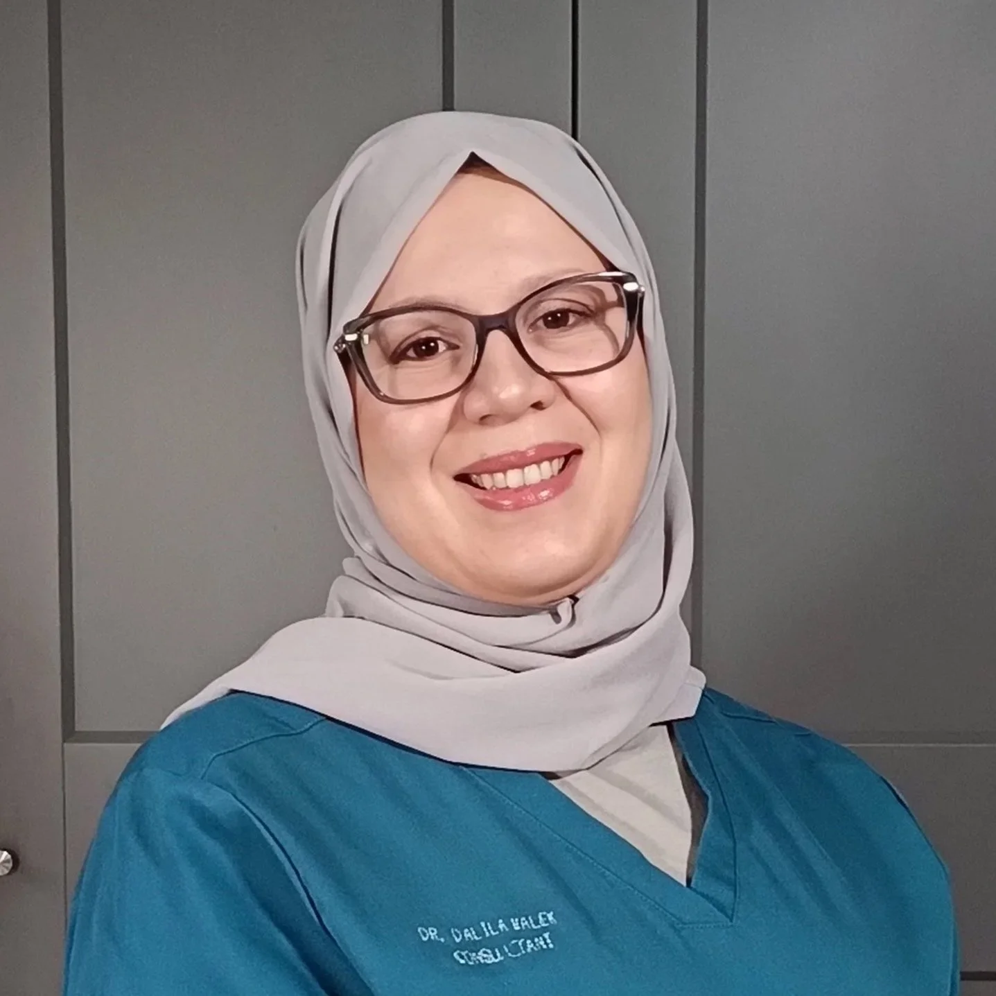 A woman wearing glasses and a white hijab with a blue medical scrub top, smiling in front of a gray background.