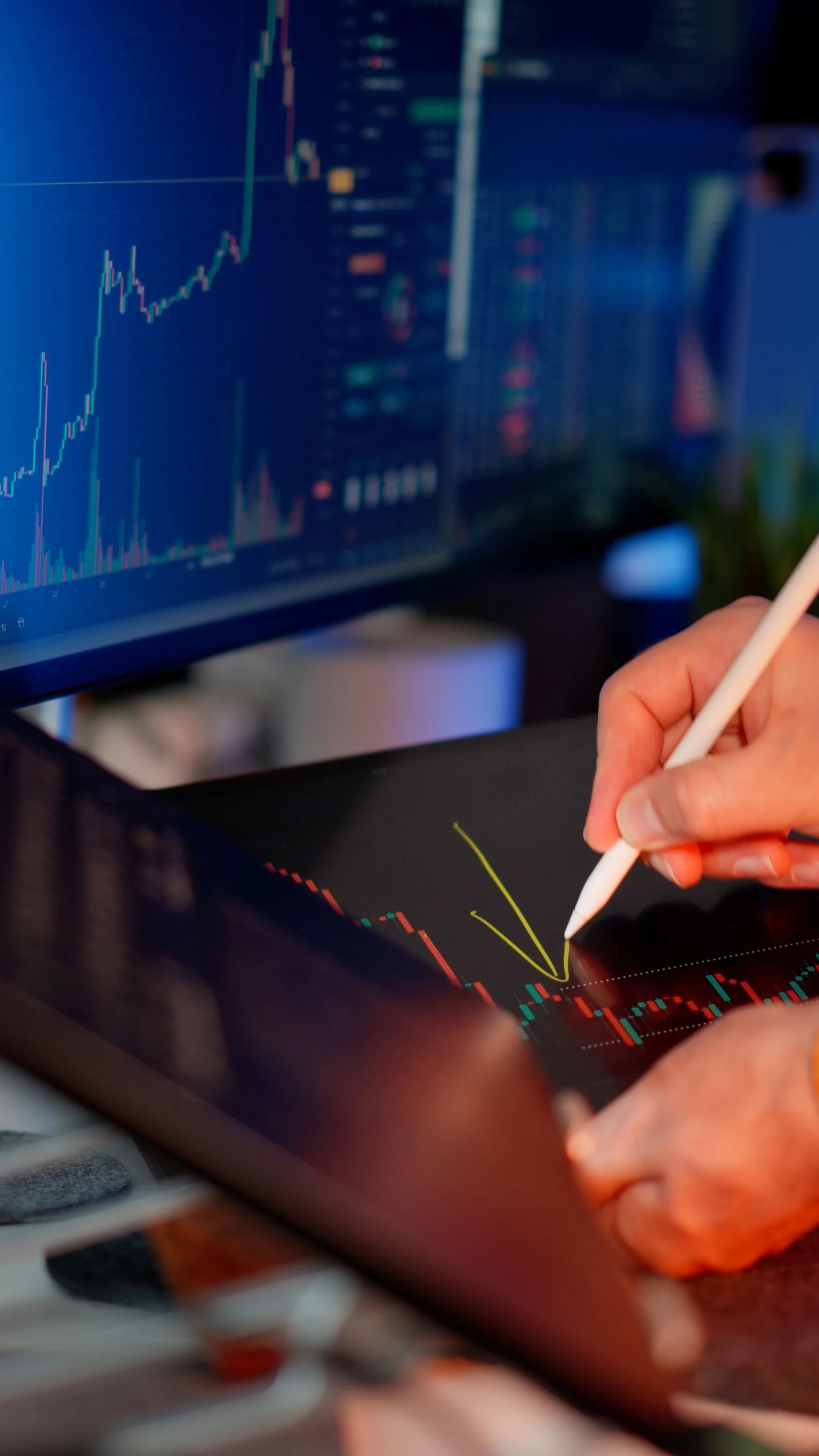 Person using a stylus on a touchscreen displaying stock market charts, with a computer screen showing financial data in the background.