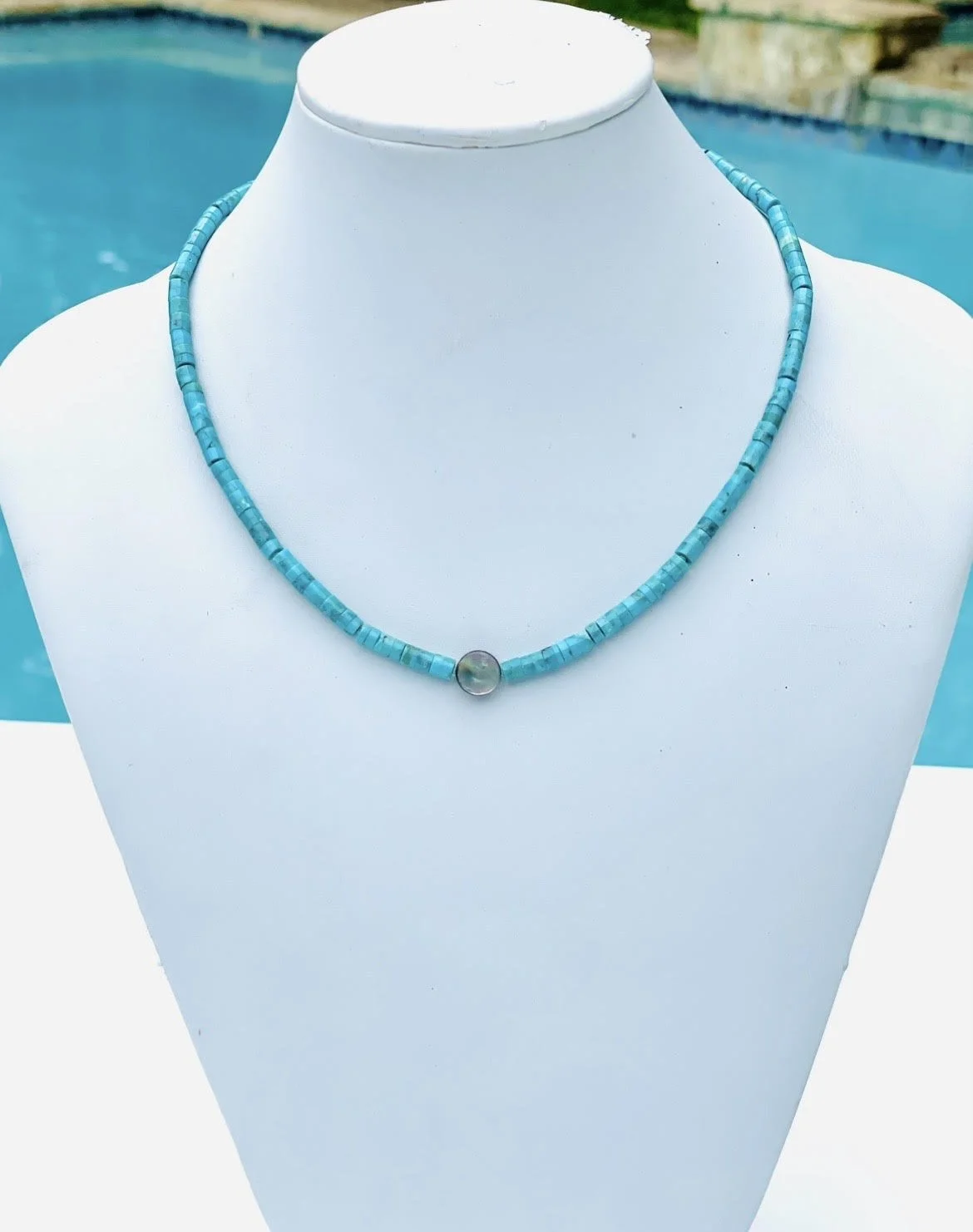 Turquoise beaded necklace with a single round clear pendant on white display bust near a swimming pool.