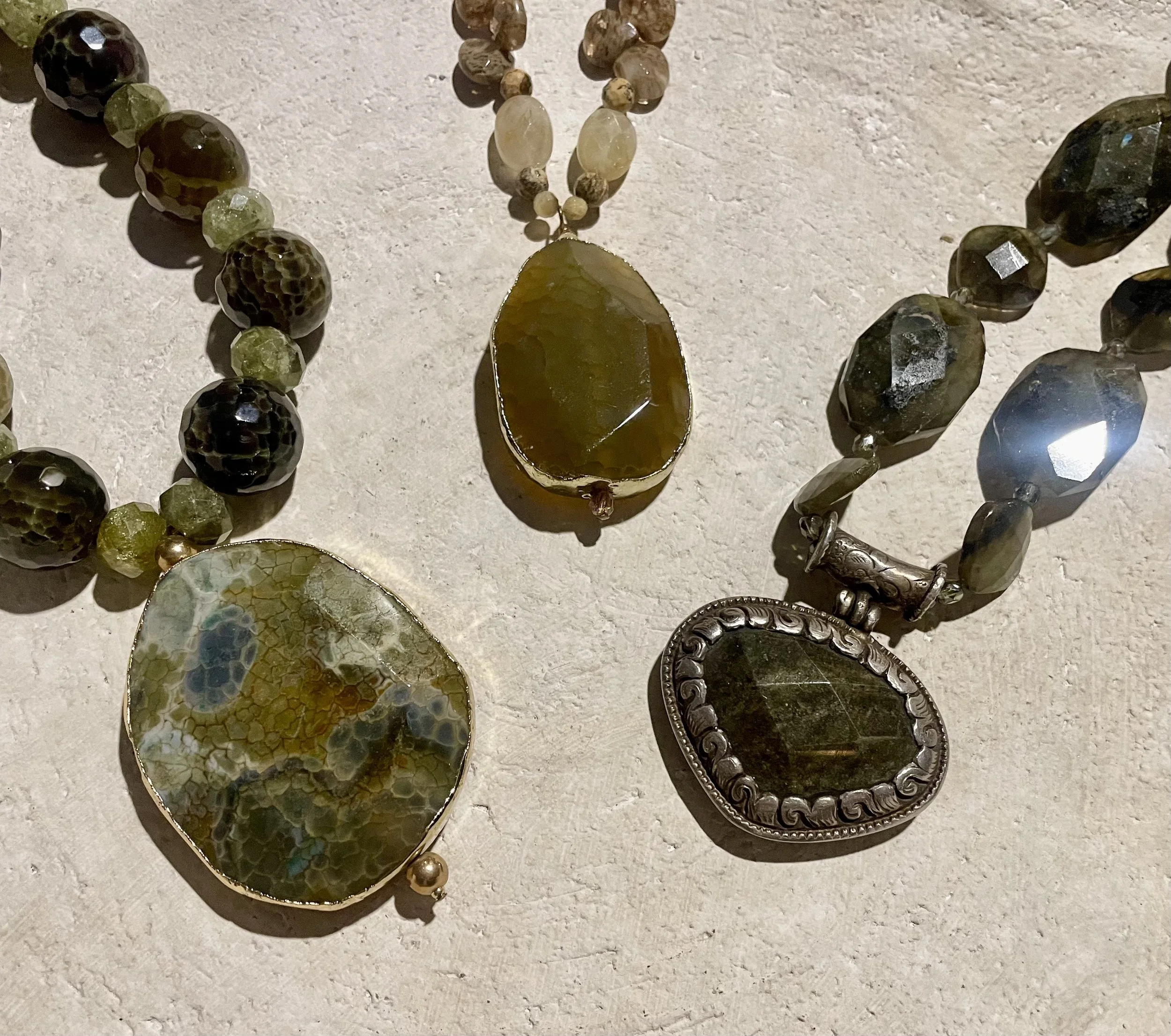 Close-up photo of various decorated jewelry pieces, including necklaces and brooches, placed on a textured surface. The jewelry features beads, stones, and metal details with different shapes, sizes, and colors.