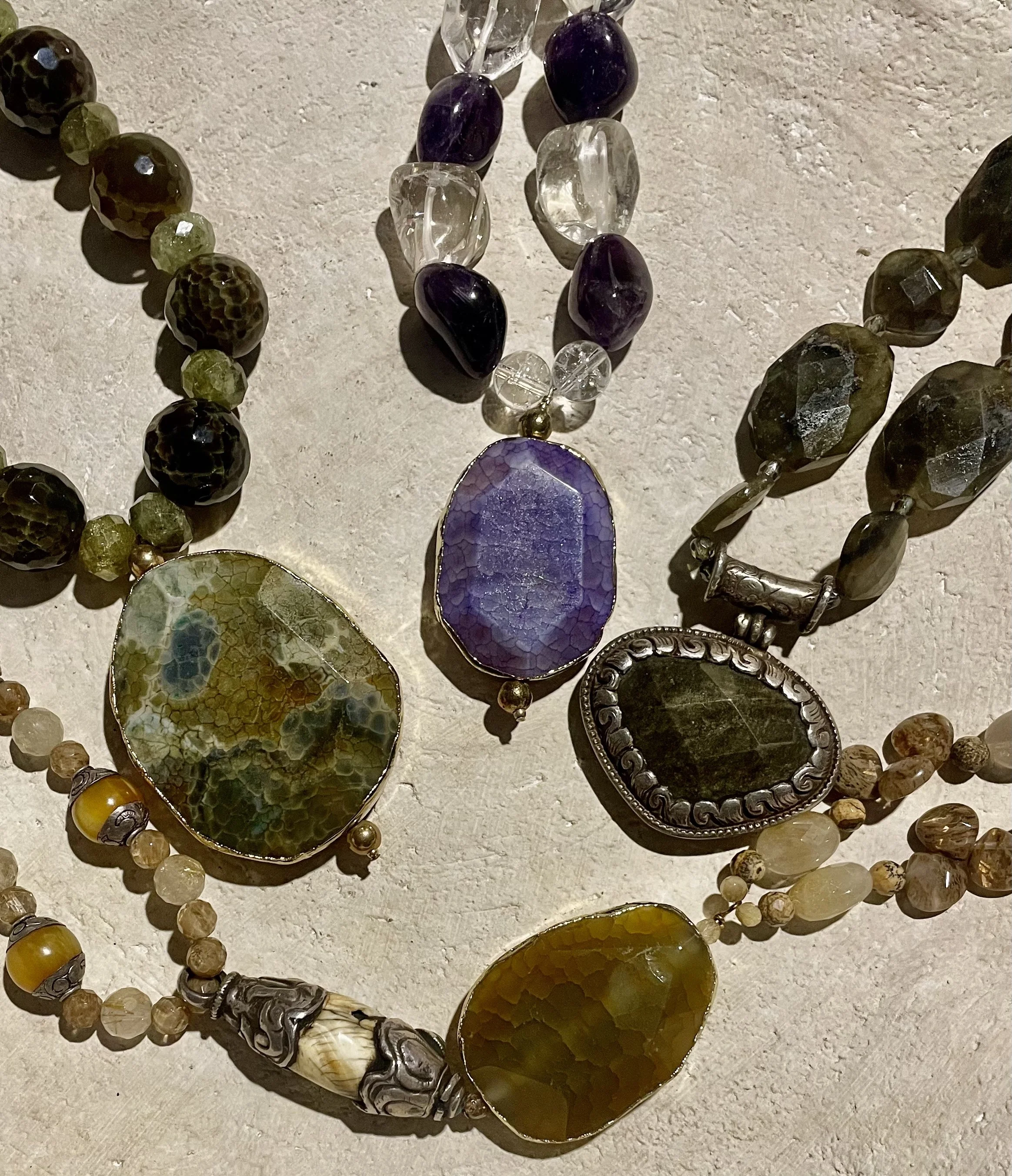 Various jewelry pieces, including beaded necklaces and large gemstone pendants, displayed on a textured surface.
