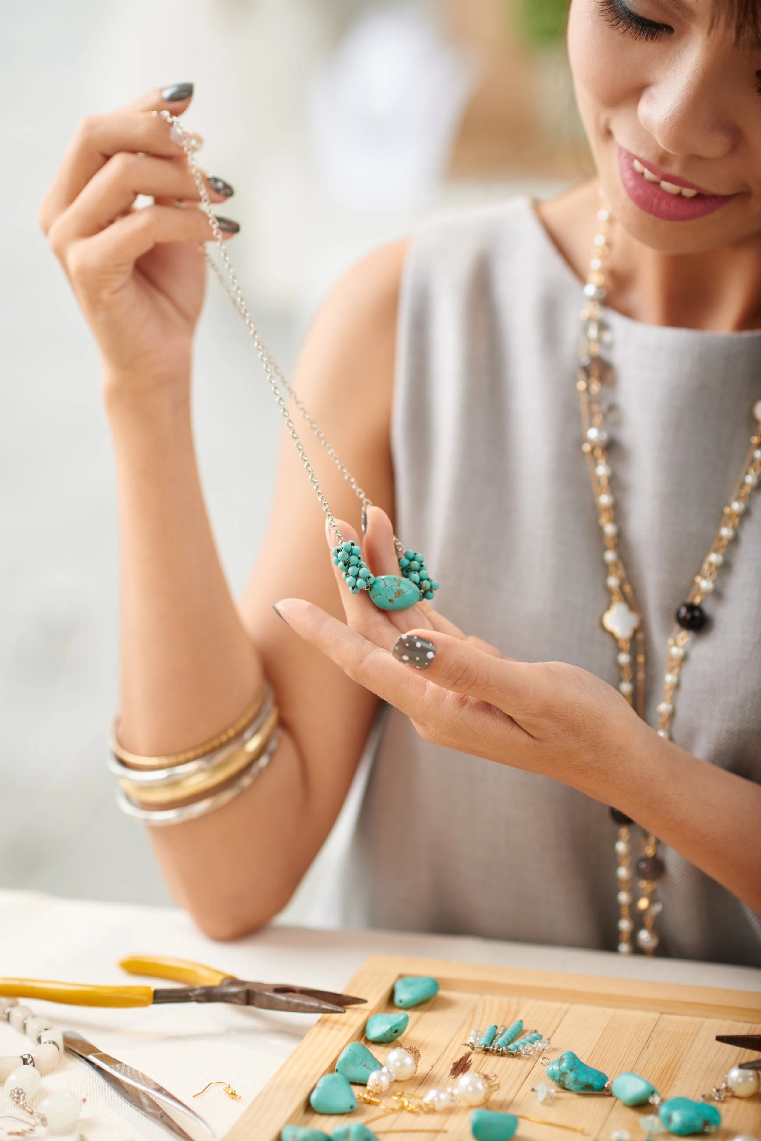 A woman wearing a sleeveless top and multiple gold bracelets is jewelry-making, handling a turquoise-colored pendant necklace. She is surrounded by various jewelry pieces, pliers, and jewelry components on a worktable with a wooden tray.