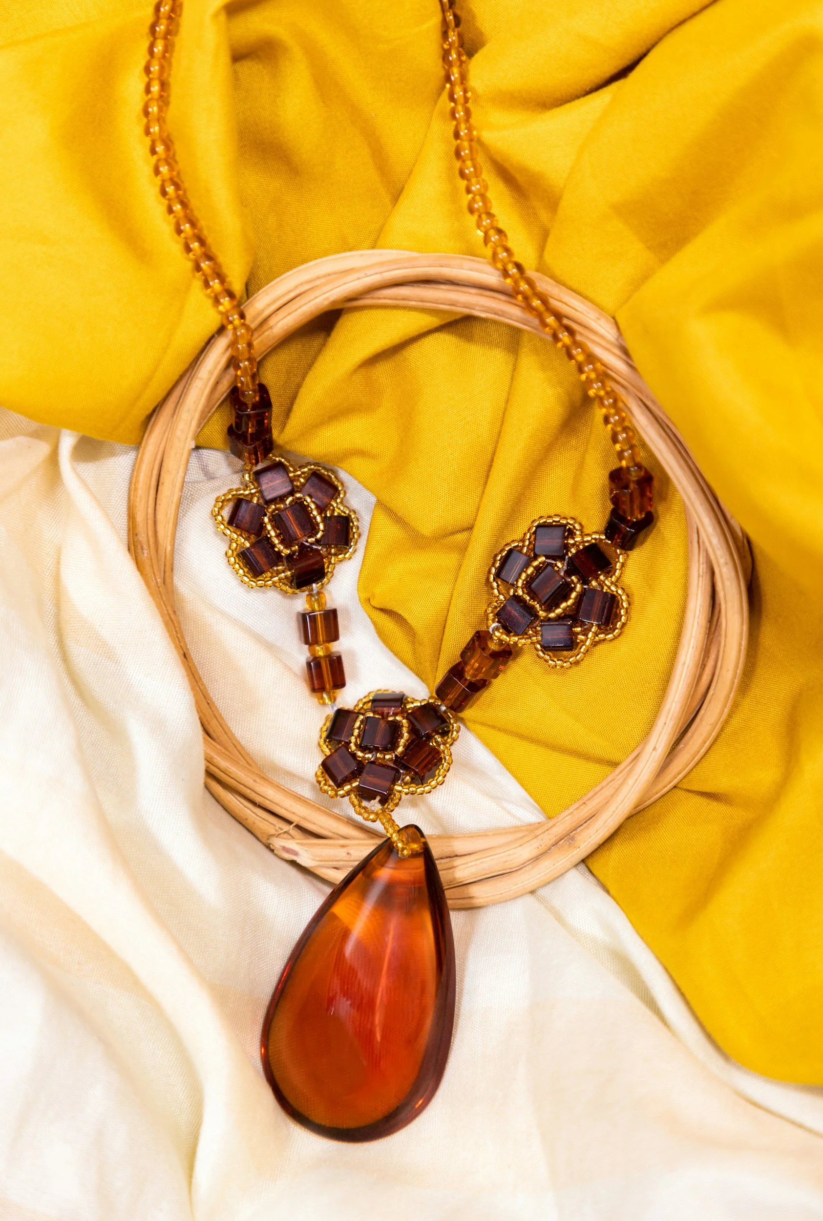 A brown and amber-colored statement necklace with a large teardrop pendant, laying on yellow and cream fabric with a wooden hoop.