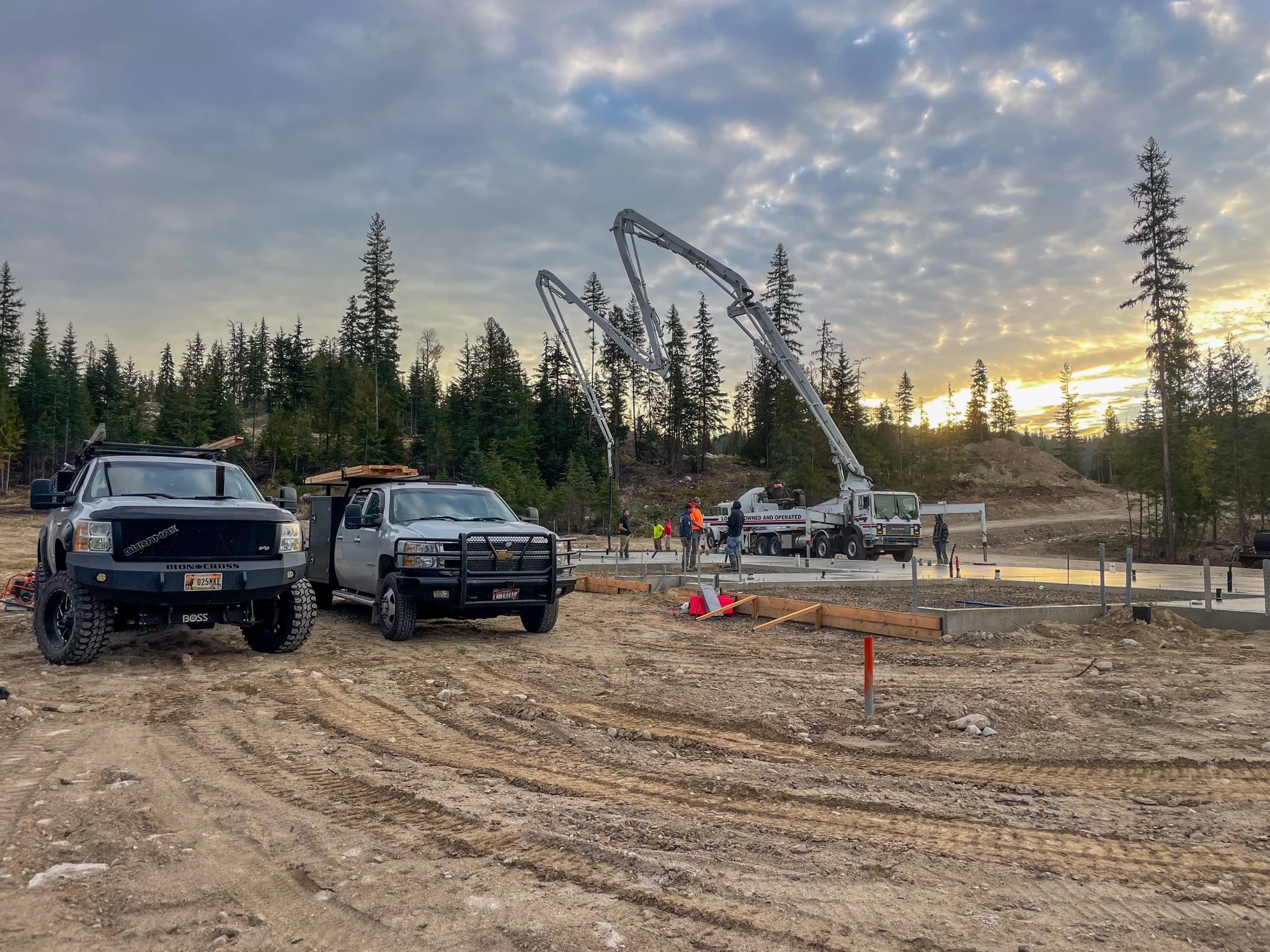 Construction site with vehicles, cranes, and people working amid trees during sunset.
